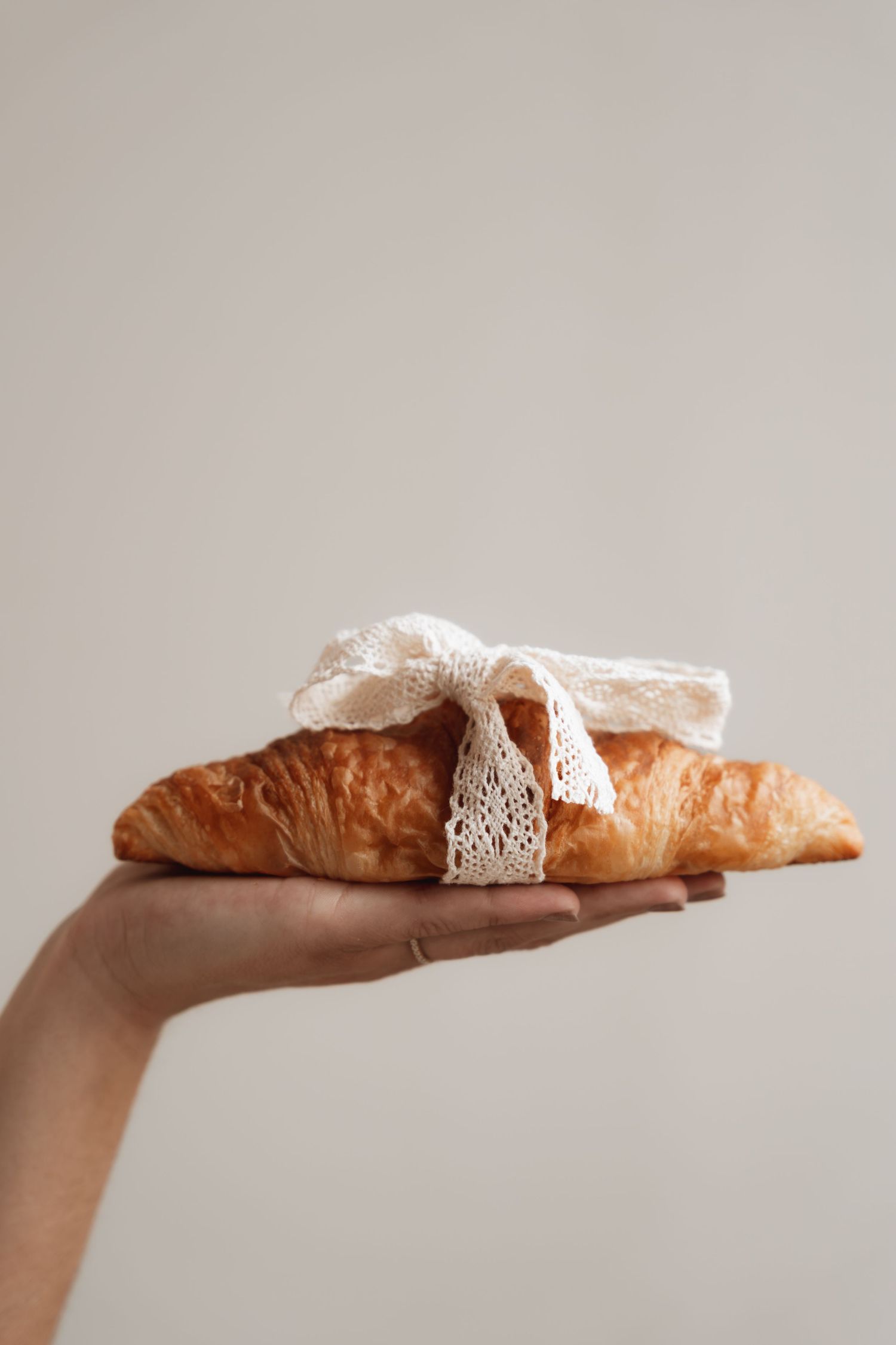 A hand holding a golden croissant tied with a delicate white lace bow against a neutral background.
