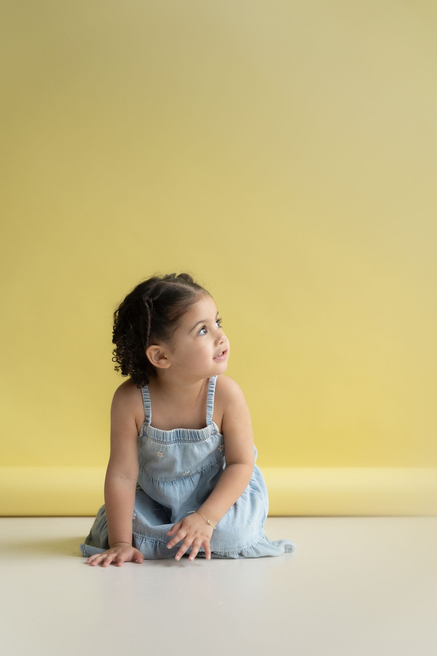 A young dancer in blue dress practices graceful poses on a yellow backdrop in a minimalist studio setting.