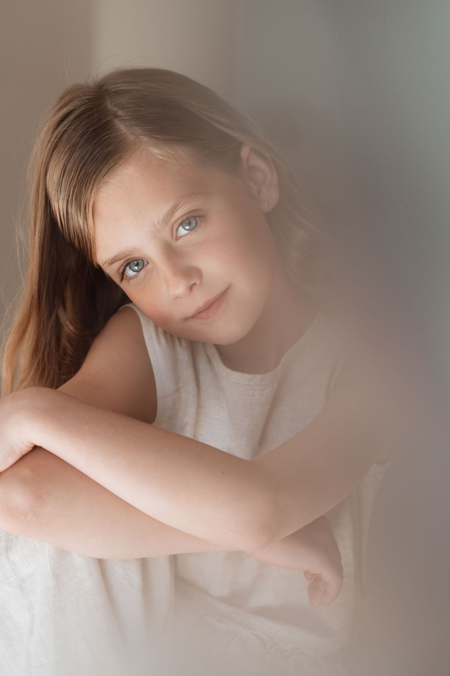 Young girl with blue eyes and long hair poses thoughtfully in a white top against a soft, dreamy background.