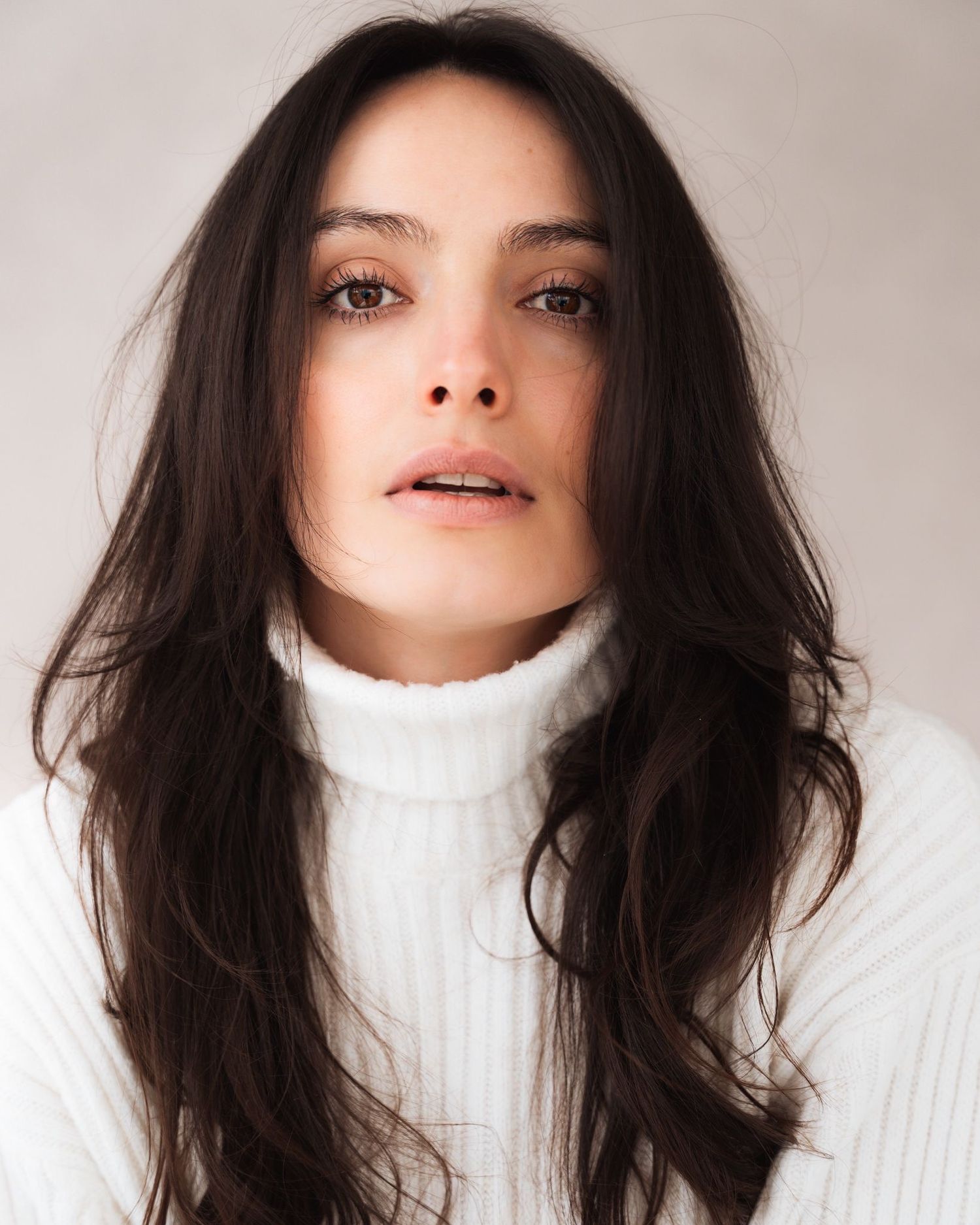 Woman in white turtleneck sweater with dark flowing hair against neutral background.