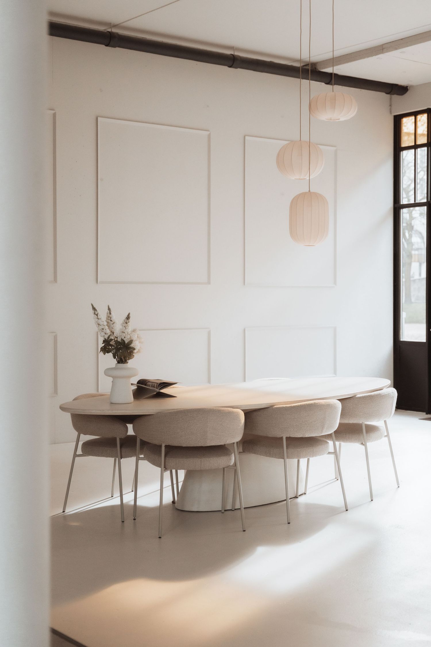 Minimalist dining room with oval table, beige chairs, white walls with panel molding, and hanging pendant lights.
