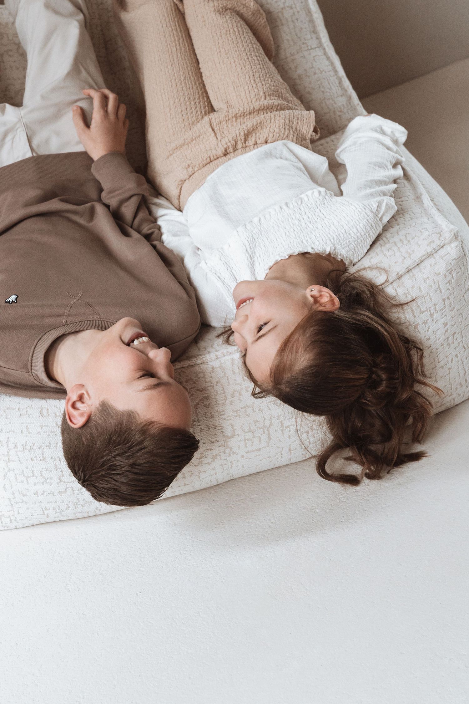 Couple lying on a couch laughing together, dressed in casual beige and white outfits in a cozy home setting.
