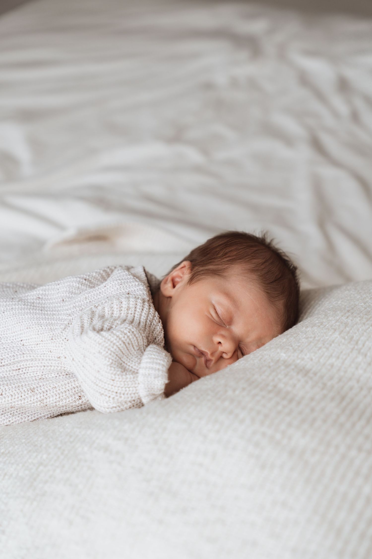 Sleeping baby in white knit sweater resting peacefully on white bedding.