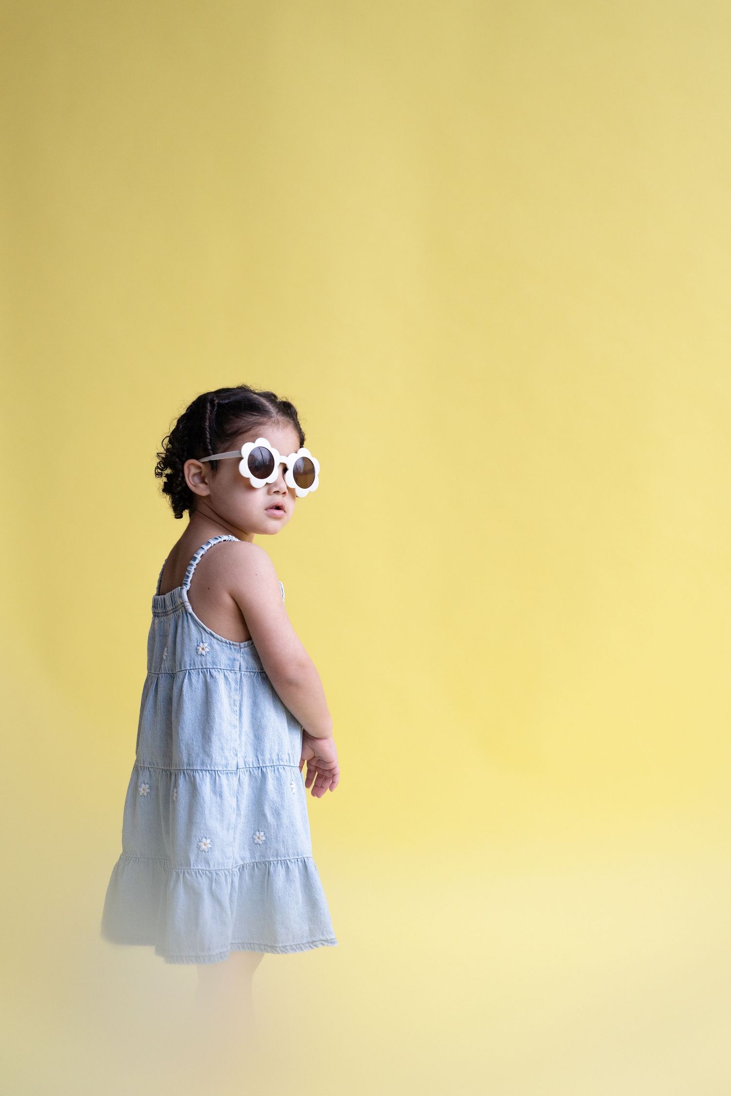 A toddler in a light blue dress and sunglasses poses against a yellow wall in a sequence of four photos.