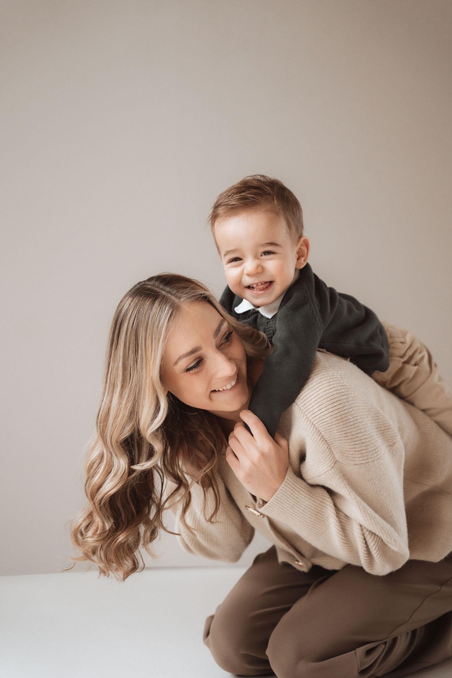 A smiling mother in a beige sweater playfully holds her happy toddler boy on her back against a neutral background.