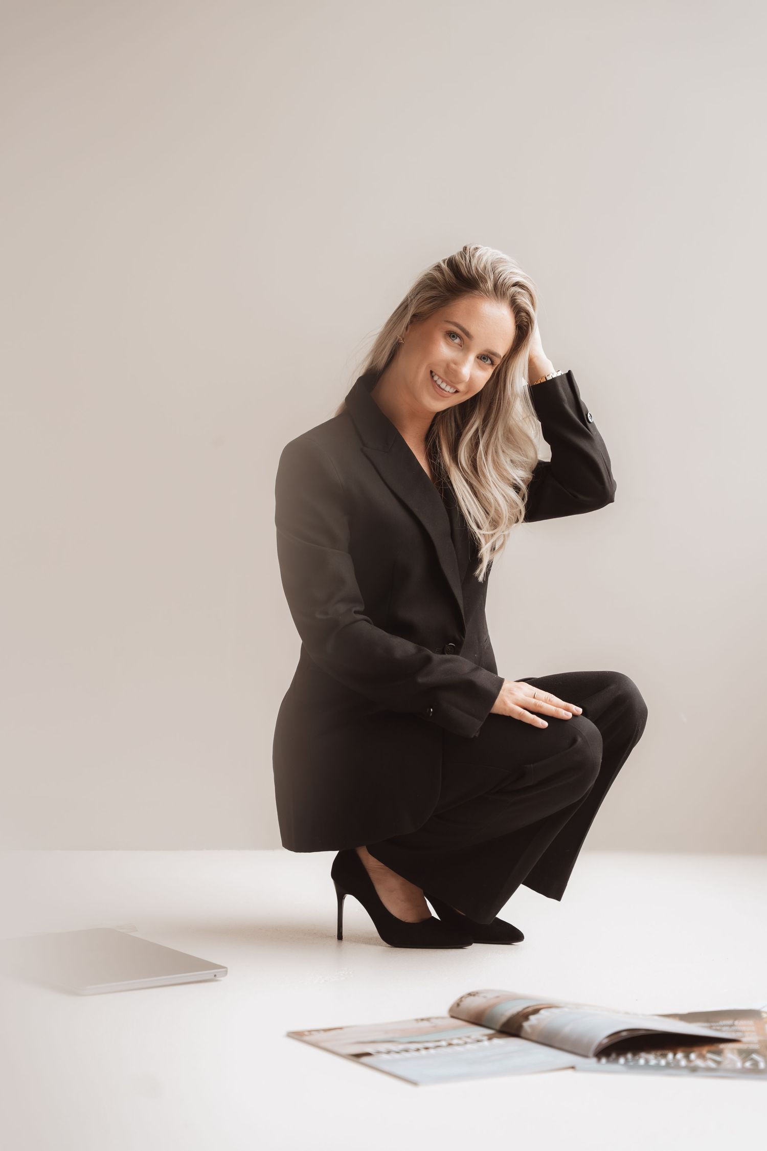 Smiling blonde woman in black business suit crouching beside a laptop and open magazine on white floor.