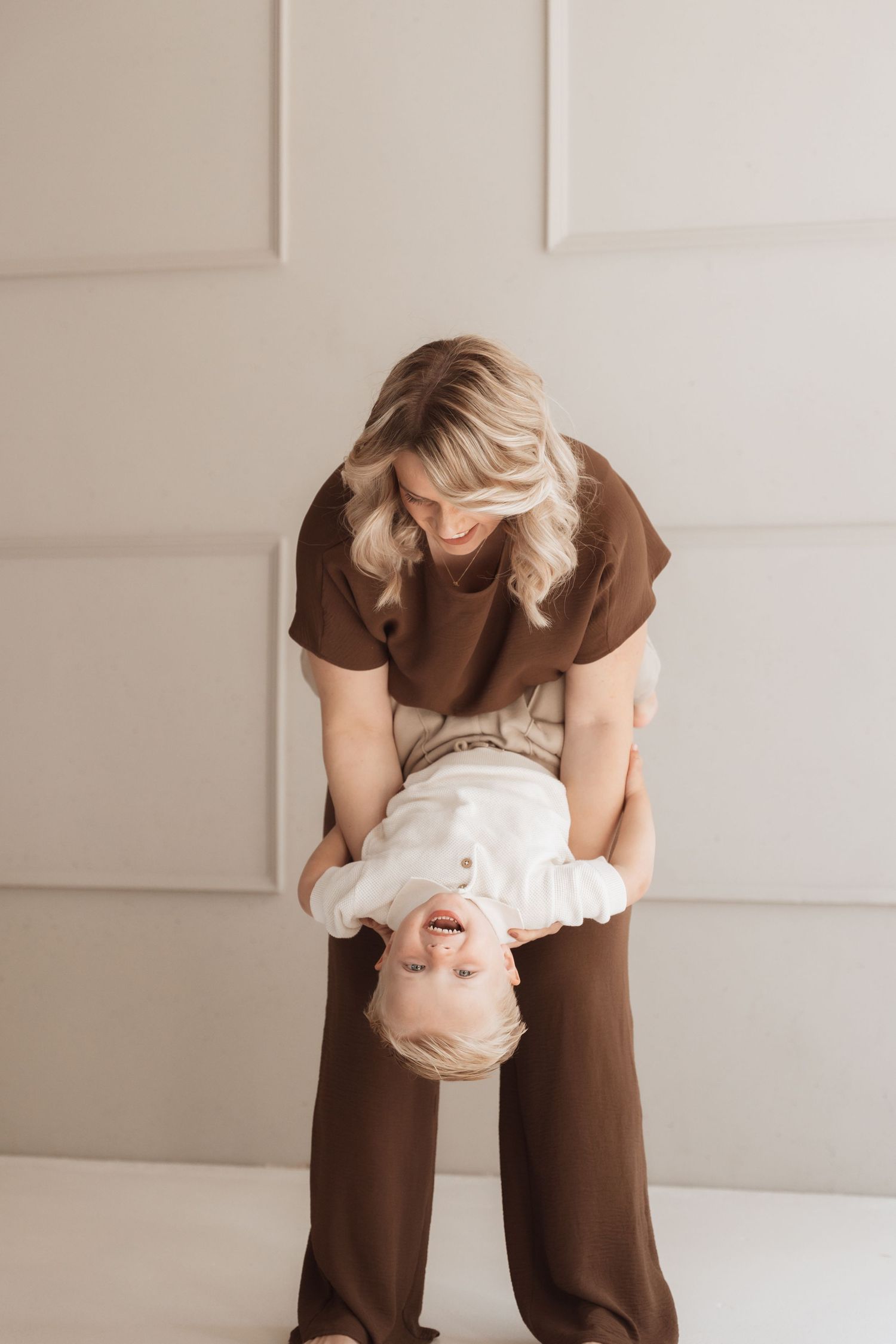 A blonde woman in brown outfit playfully holds a laughing toddler upside down in a bright, neutral-toned room.
