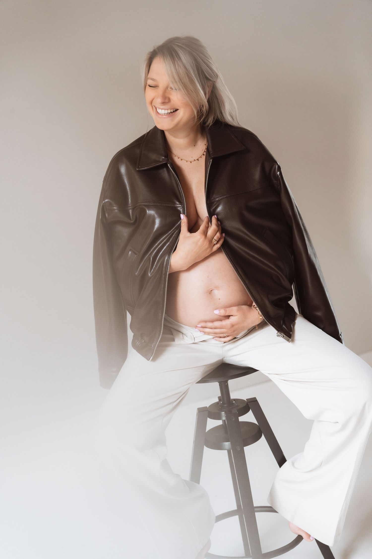 Smiling pregnant woman with gray hair wearing a brown leather jacket and white pants, sitting on a stool showing her baby bump.