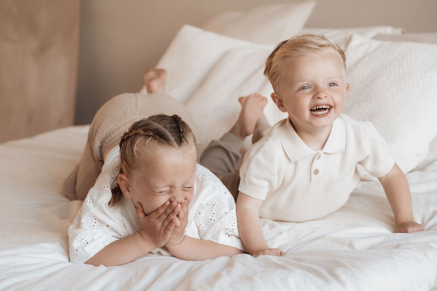 Two laughing toddlers lying on a white bed, a girl covering her mouth and a boy smiling brightly.