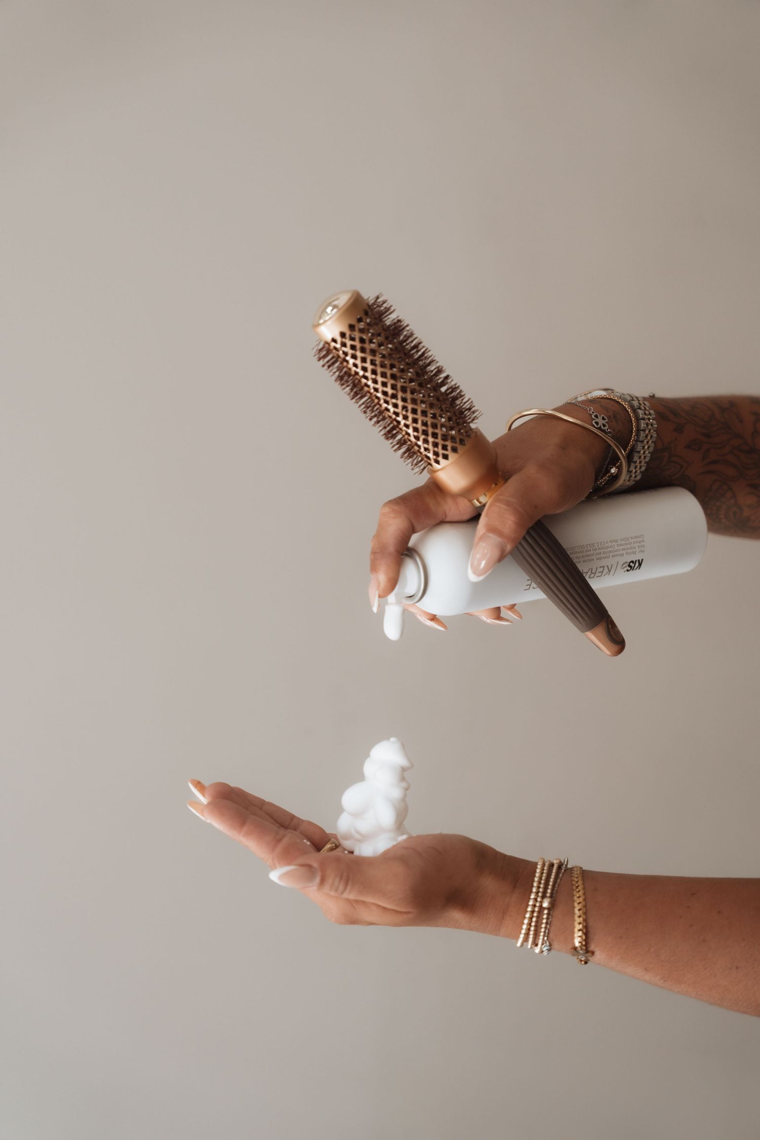 Hands dispensing hair mousse foam while holding a gold round brush, wearing gold bracelets, against neutral background.