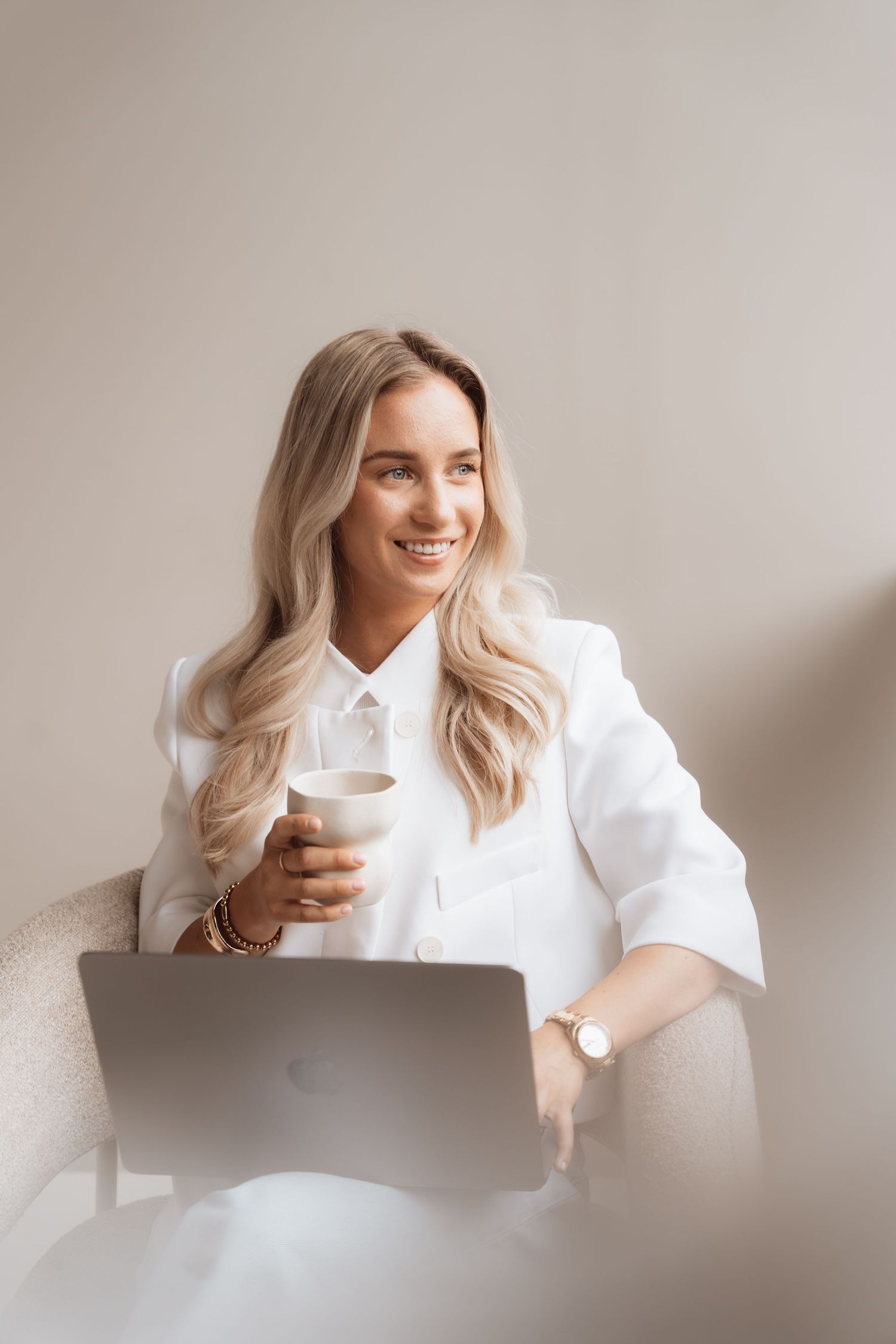 Smiling blonde woman in white blazer holding coffee cup while working on MacBook laptop in bright minimalist setting.
