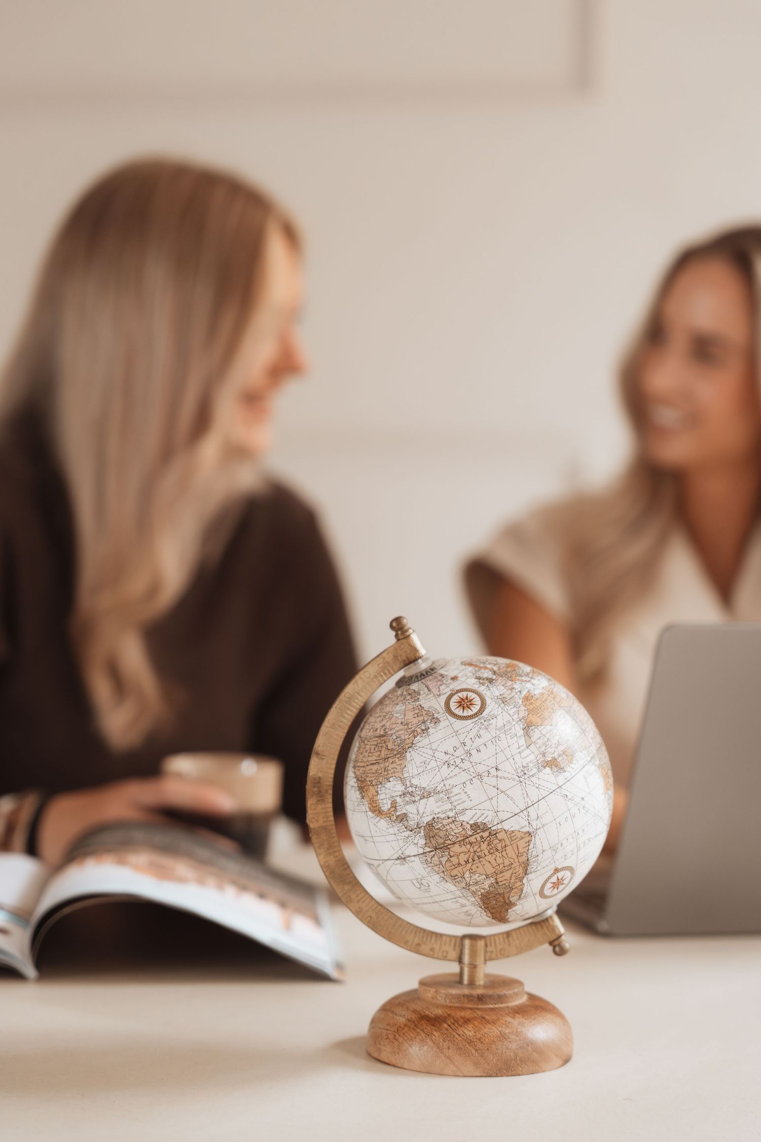 Decorative vintage globe on desk with two women discussing travel plans using laptop and magazine in background.