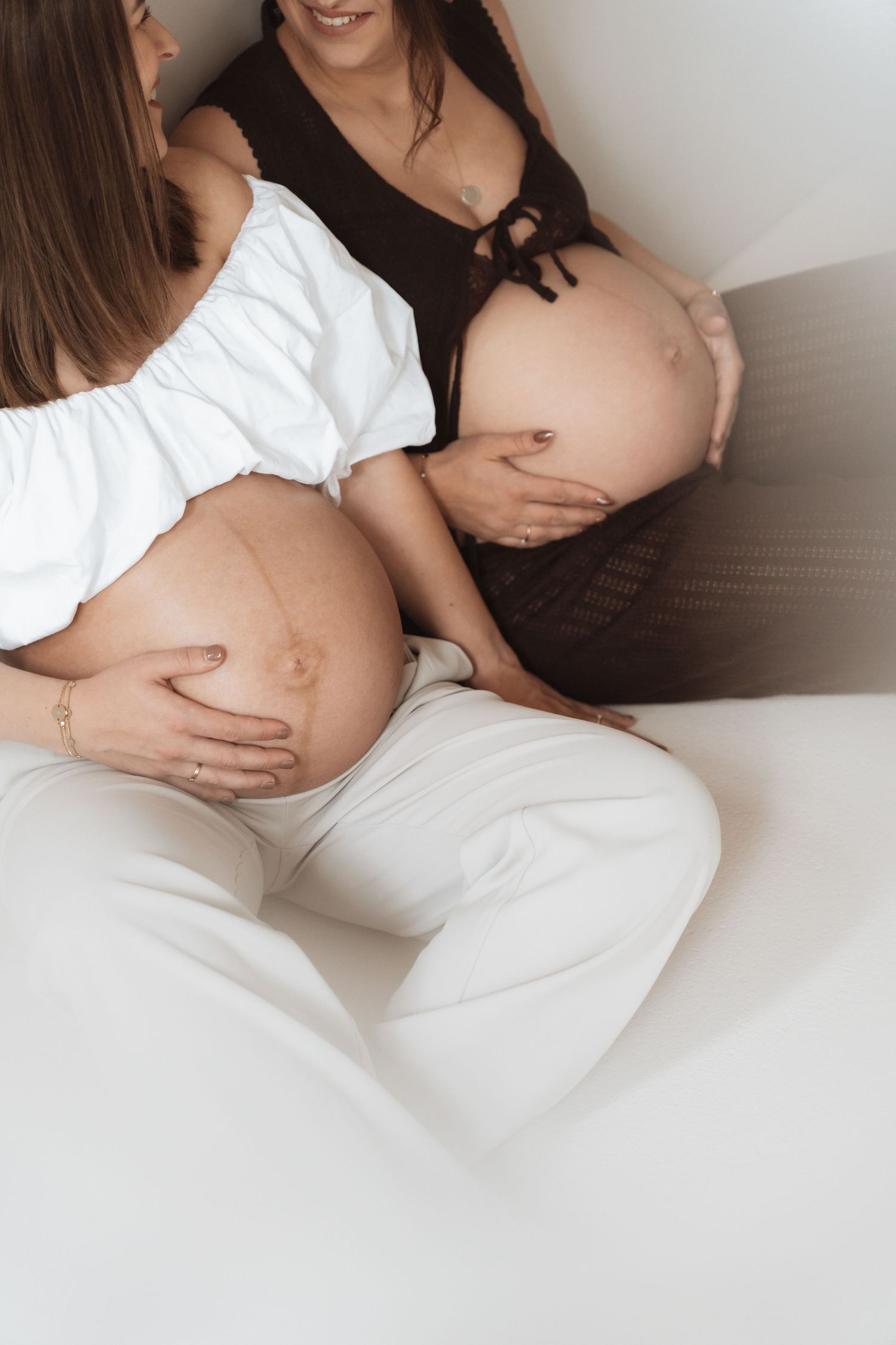 Two pregnant women sitting together, cradling their baby bumps, wearing white and dark outfits in a bright room.