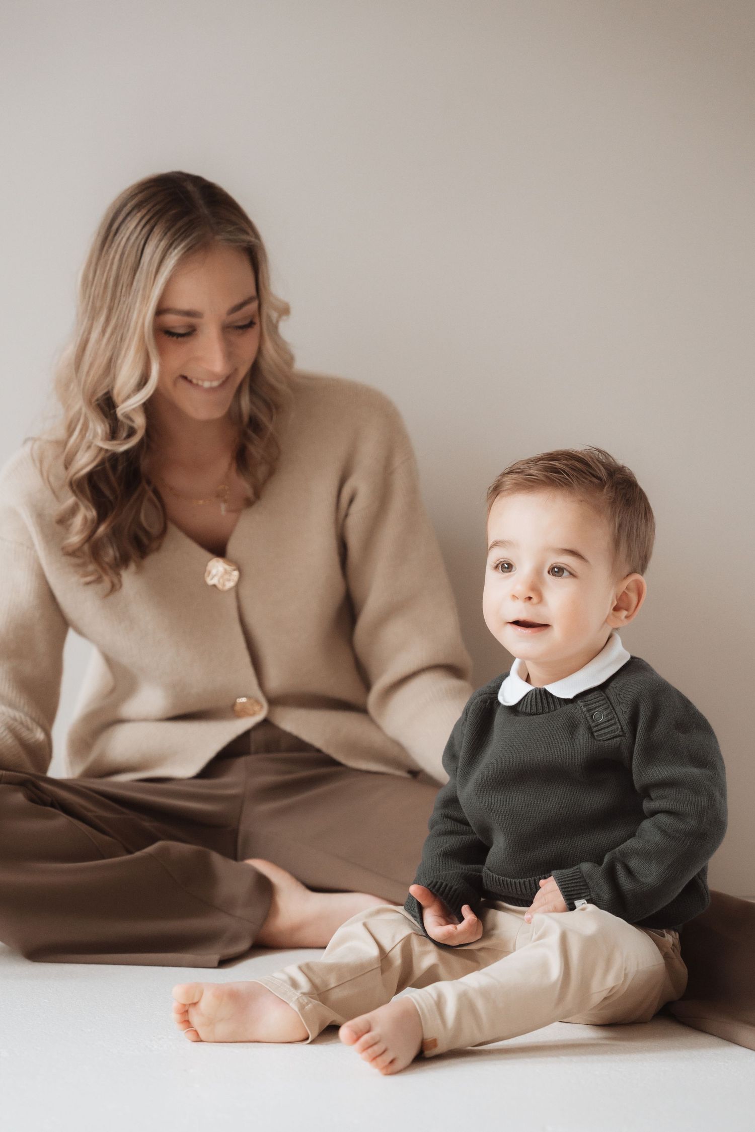 A cozy portrait of a parent and young child sitting together in neutral-toned clothing against a beige wall.