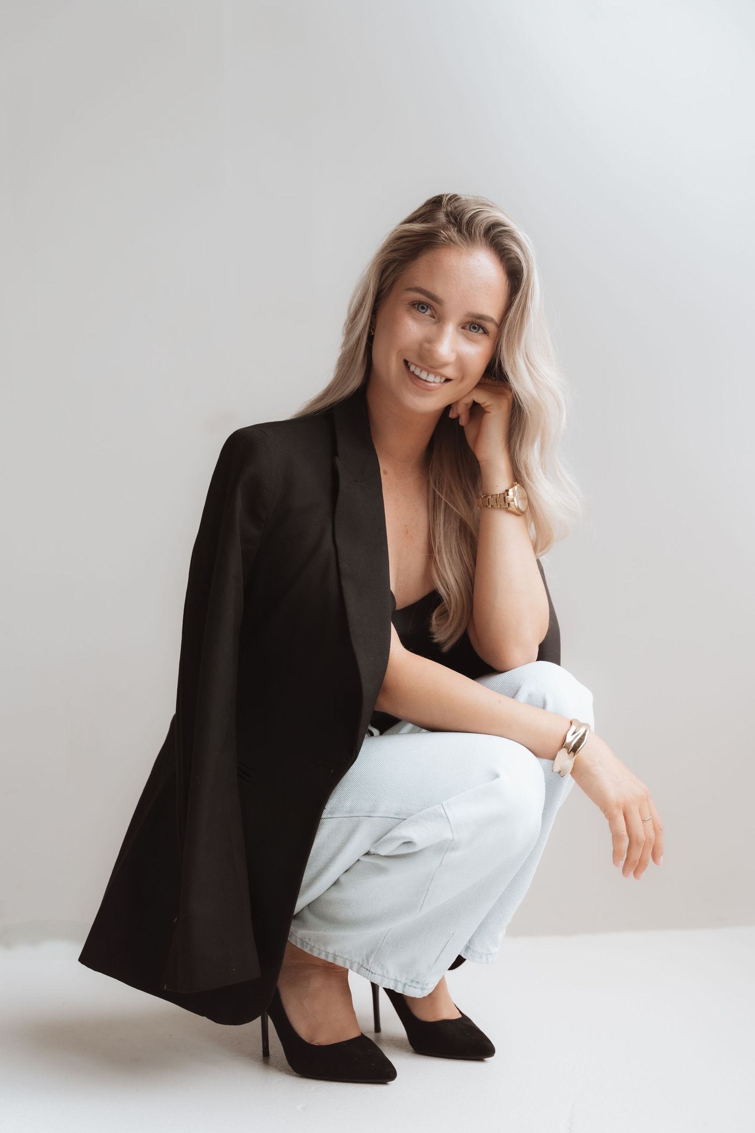Smiling blonde woman in black blazer, light jeans, and heels crouching against a white background.
