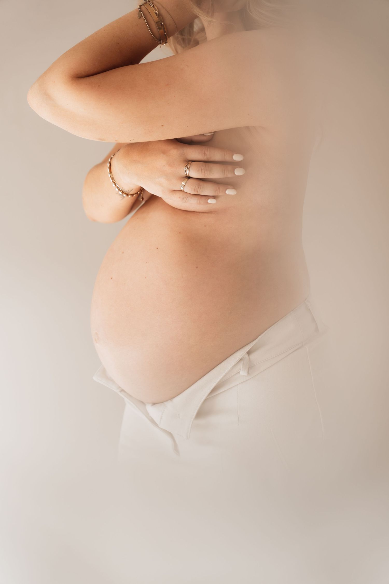 Side profile of a pregnant woman in white underwear, arms crossed over chest, wearing gold bracelets and rings.