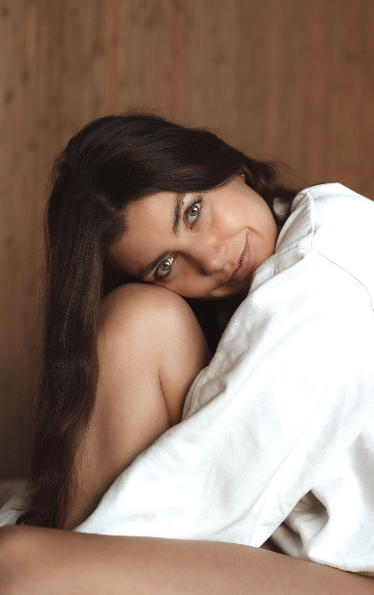 Figure wrapped in white sheets poses intimately against wooden background.