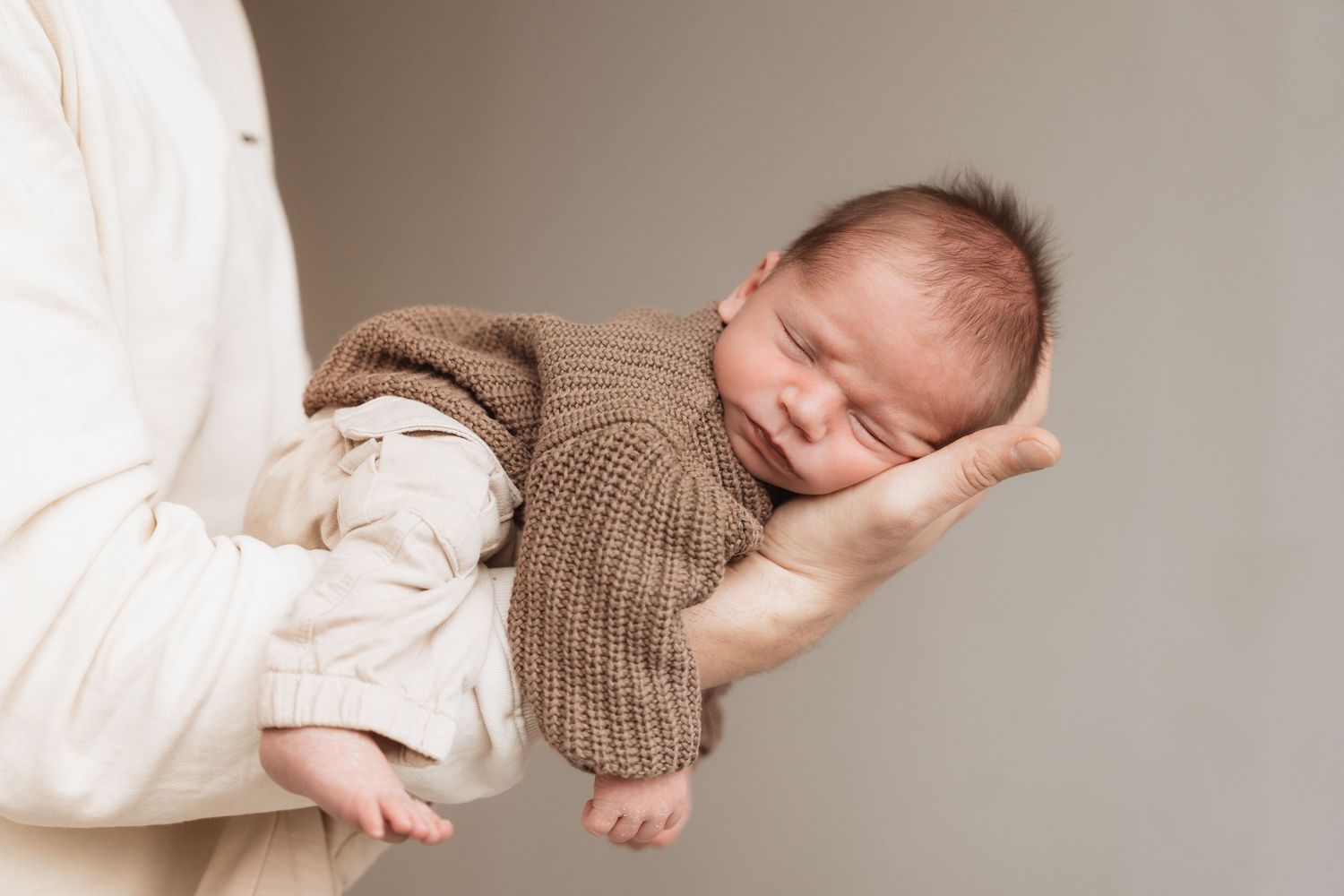 Newborn baby wearing a soft brown knitted sweater lies peacefully on white bedding.