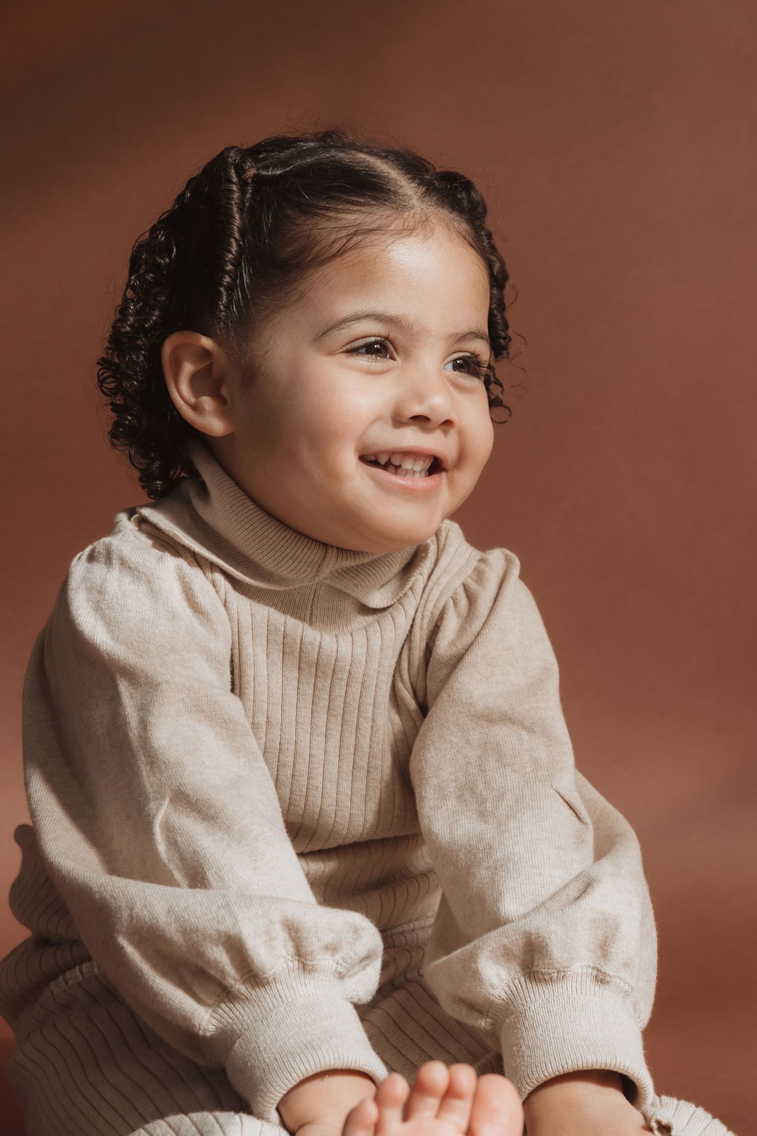A smiling toddler girl with curly pigtails wearing a beige ribbed turtleneck sweater set against a warm brown background.