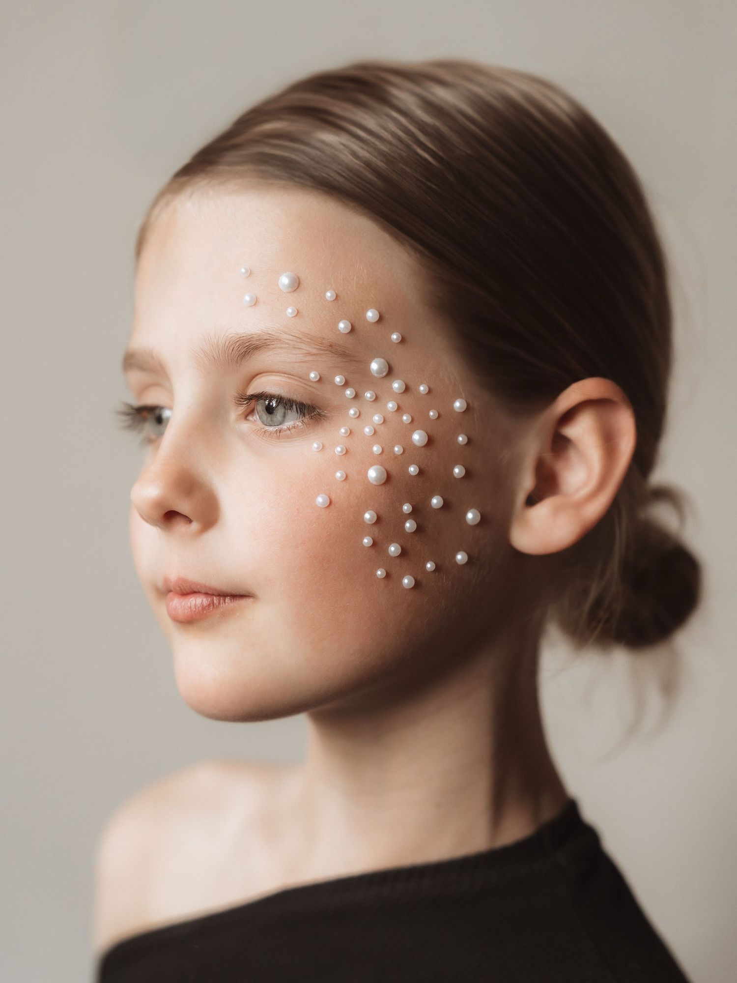 Young girl with pearl gem stickers scattered across her cheek and brow, wearing a black off-shoulder top, soft studio portrait.