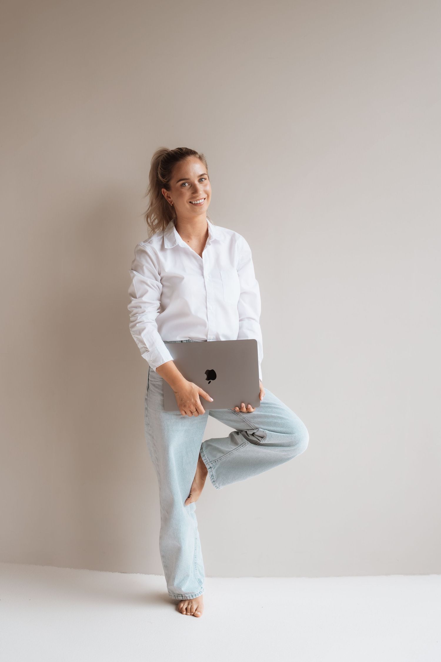 Smiling woman in white shirt and blue pants balancing on one leg while holding a laptop against a neutral background.