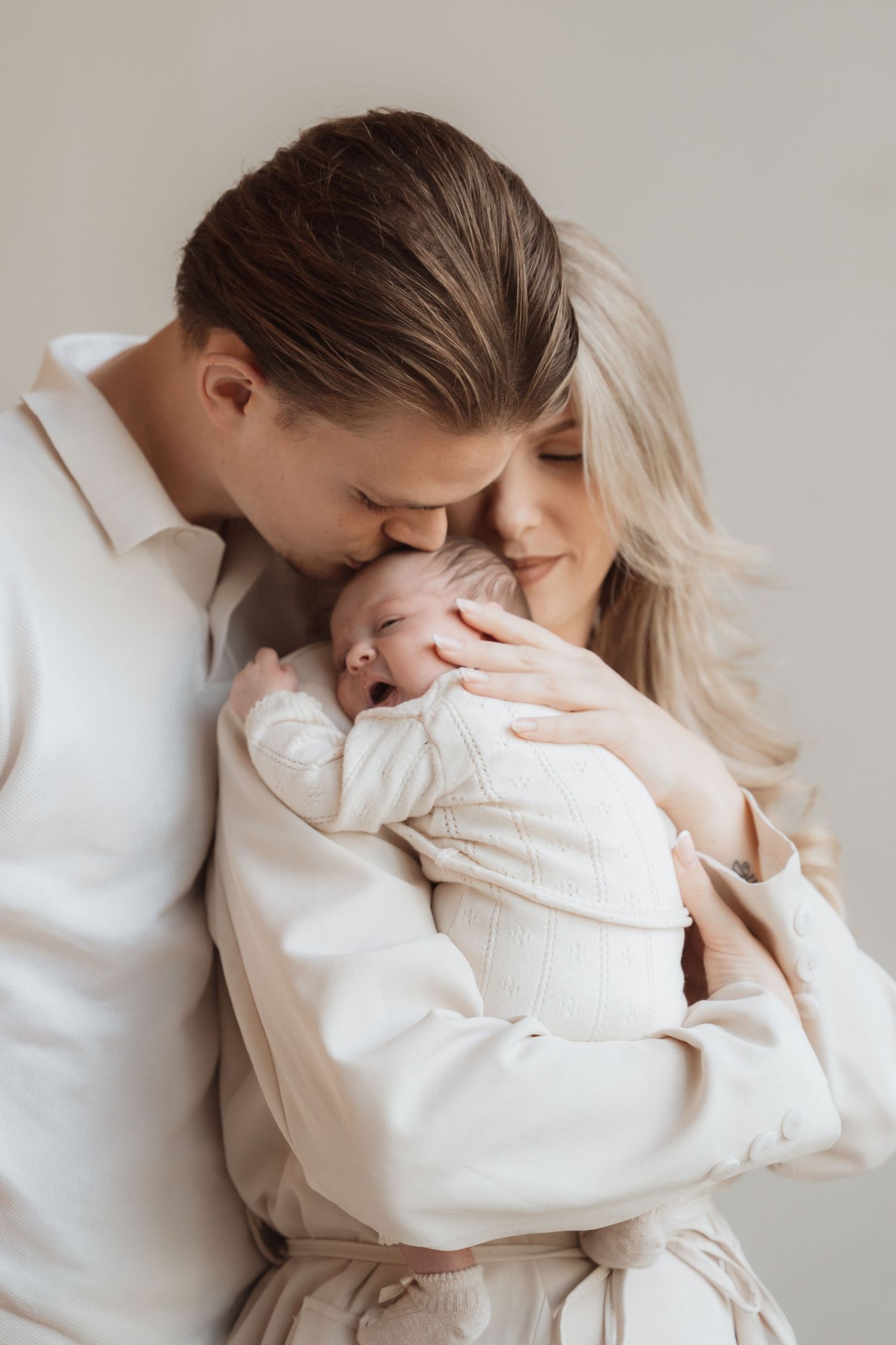 Tender embrace between parent and baby both dressed in white against neutral background.