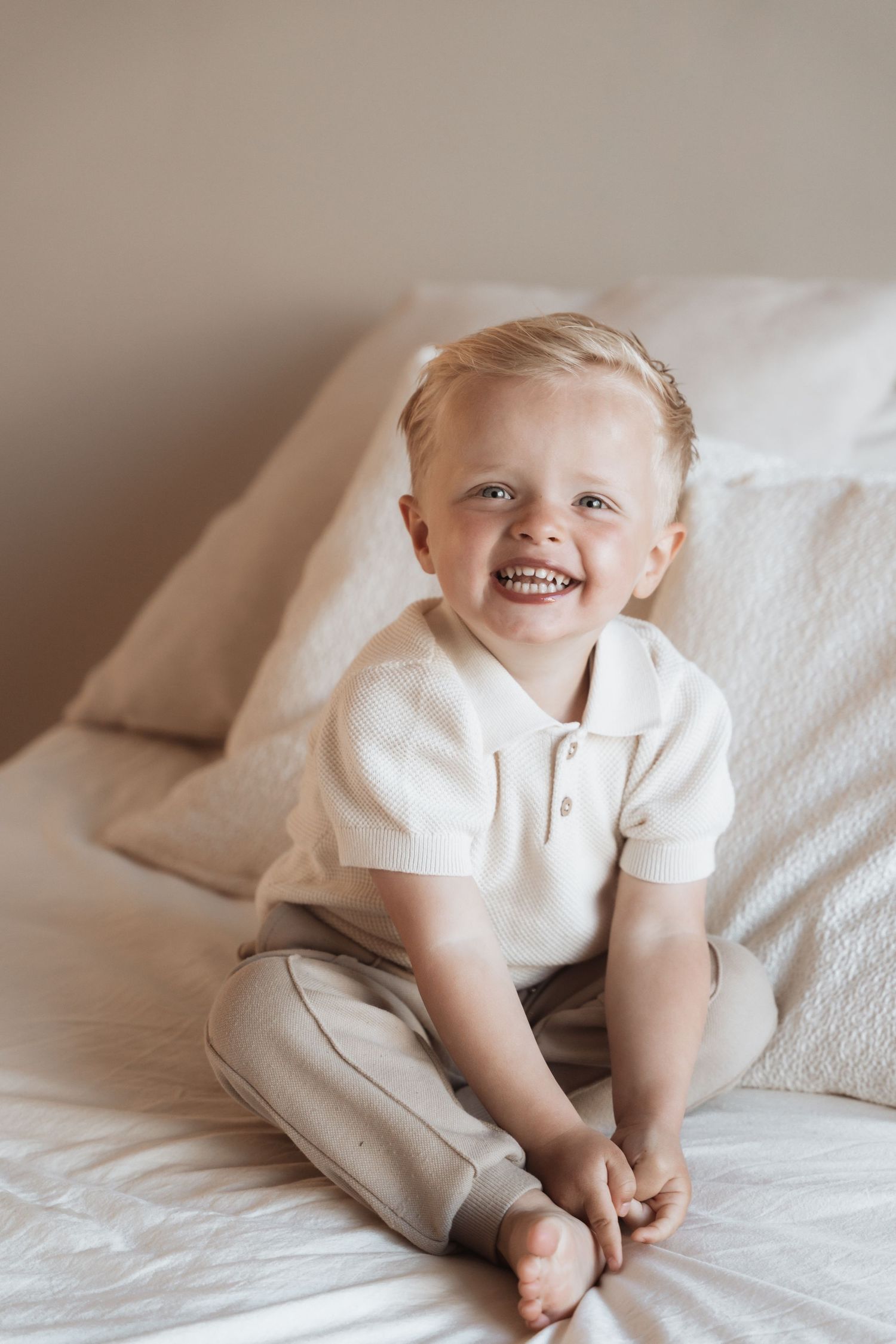 Smiling toddler boy with blonde hair sitting on a bed wearing a white polo shirt and beige pants.