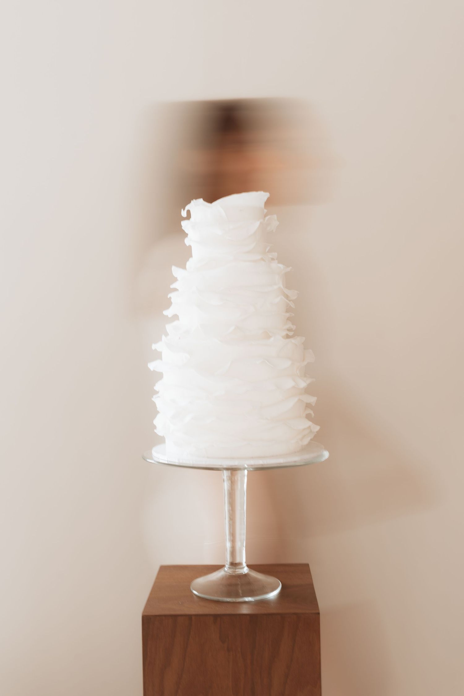 Elegant white ruffled wedding cake on a glass pedestal stand displayed on a wooden block against a neutral wall.