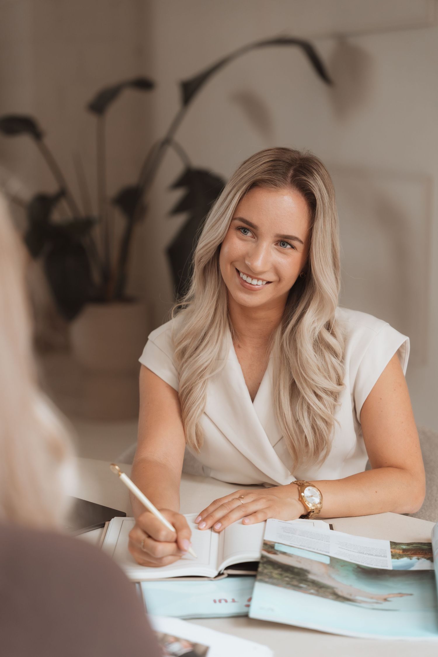 Smiling blonde woman in white blazer writing in notebook at desk with magazines in bright office.