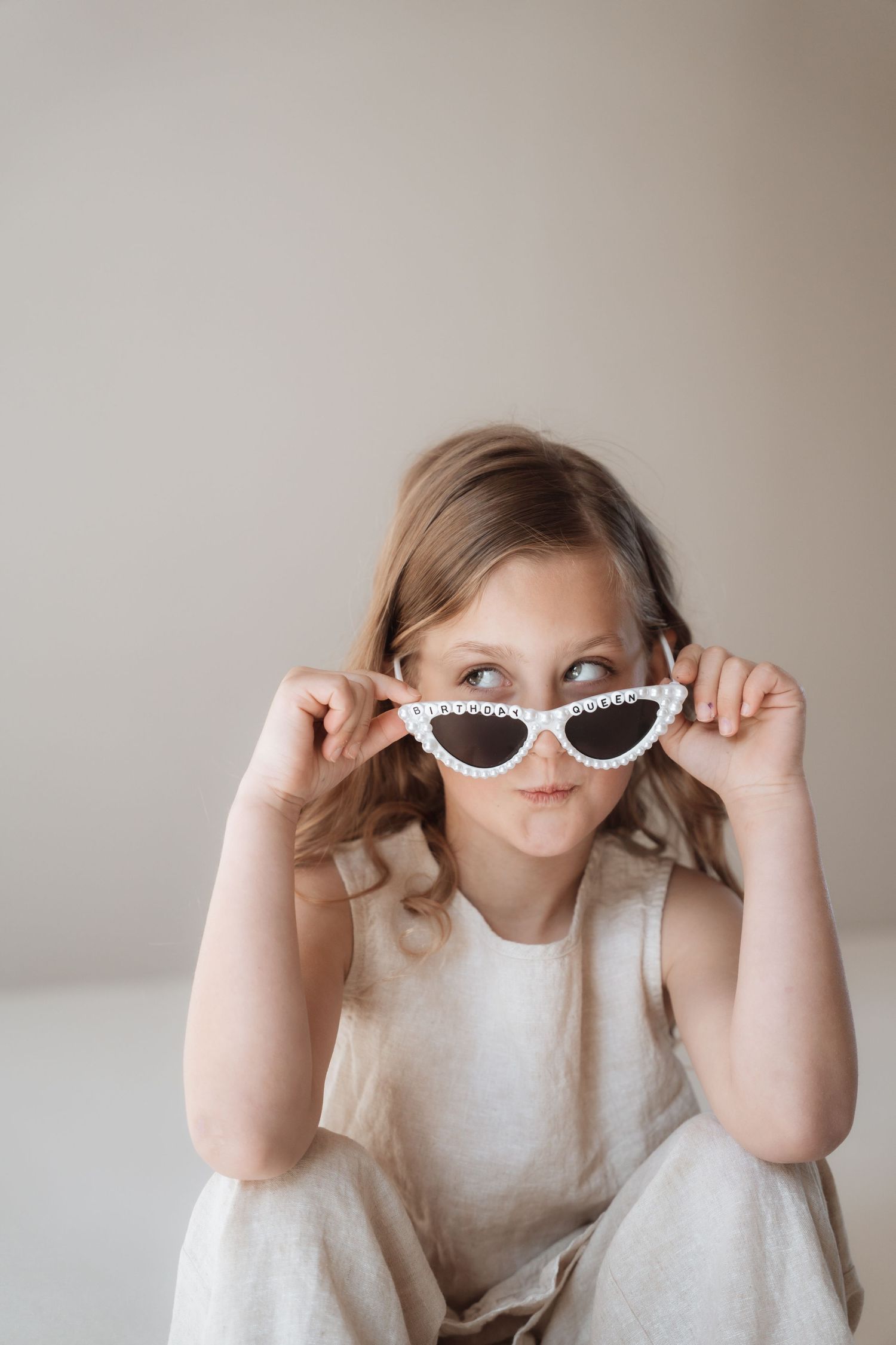 Young girl in beige dress playfully peering over white cat-eye sunglasses with pearl details against neutral background.
