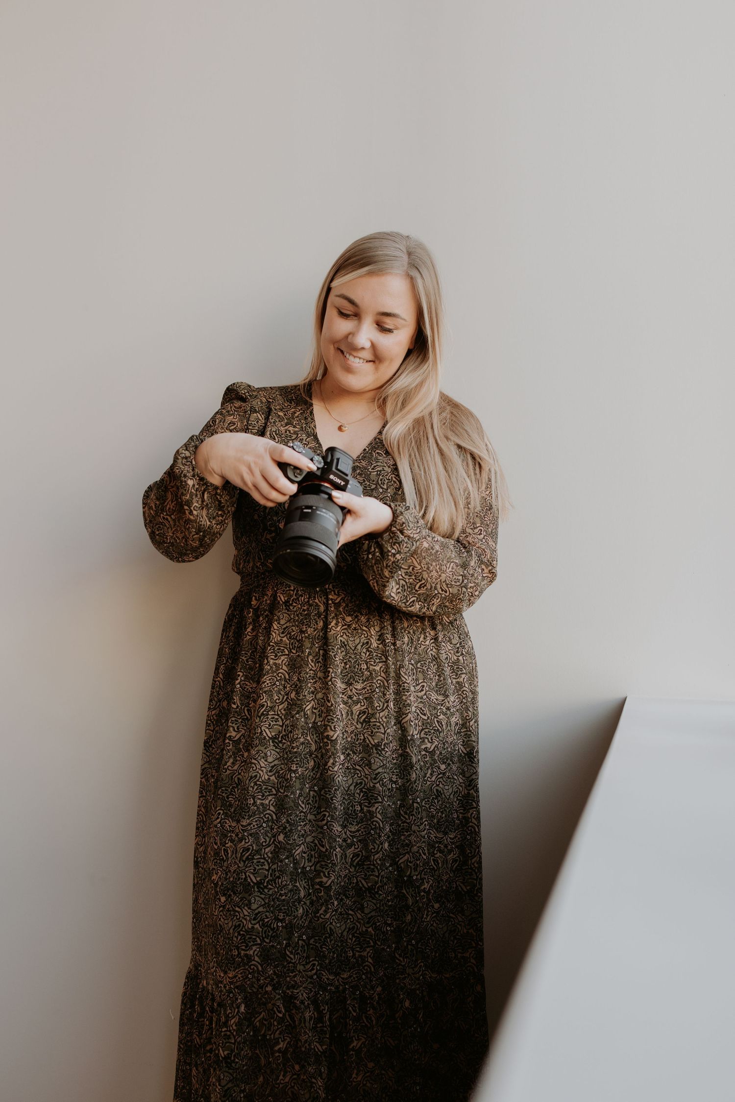 A person wearing a long brown floral dress holds a camera while smiling against a gray wall.