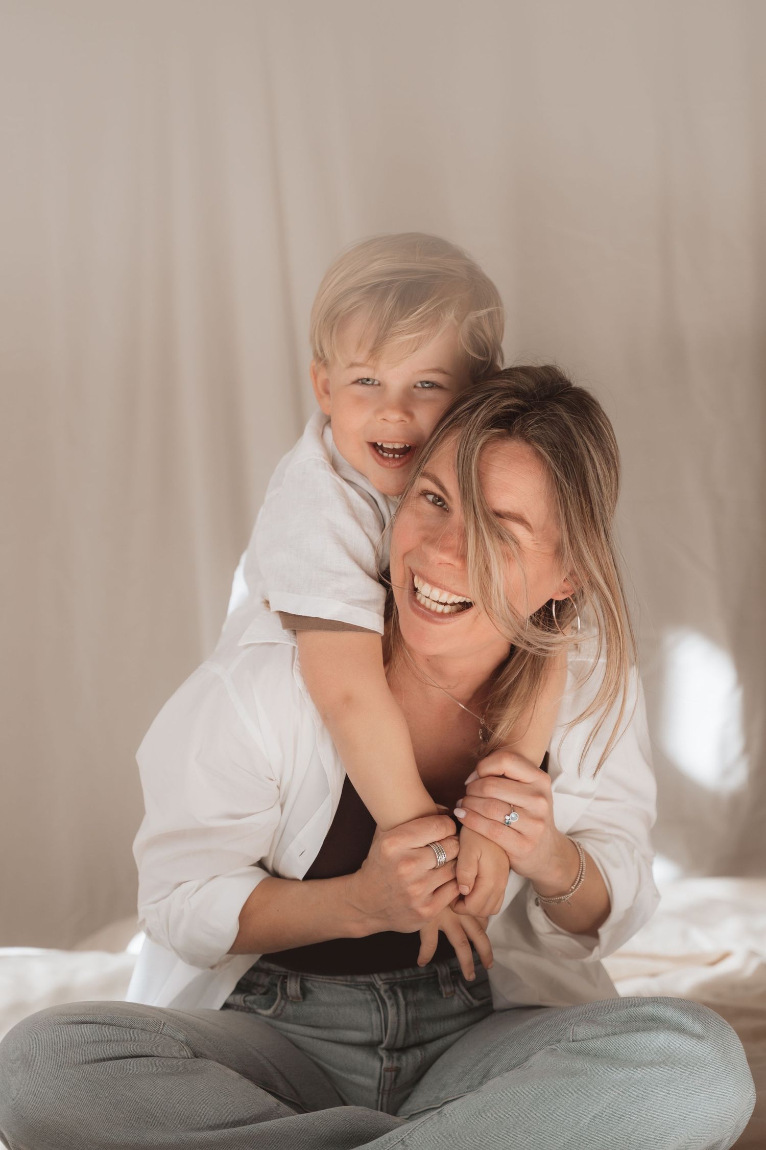 A joyful mother and young blonde son share a warm hug, both laughing, dressed in white shirts and jeans.
