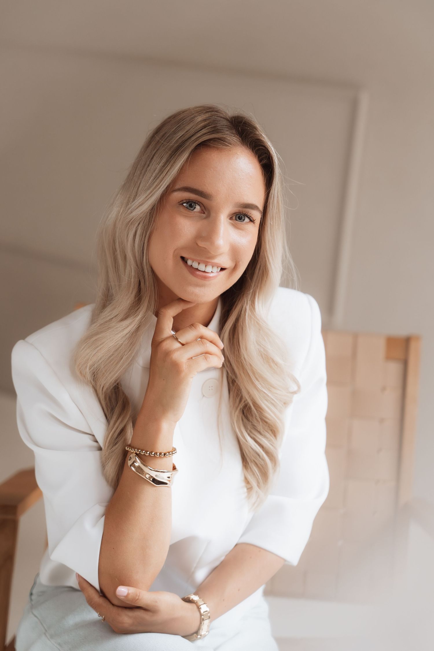 Smiling blonde woman in white blazer posing with hand on chin, wearing gold bracelets in a bright modern interior.