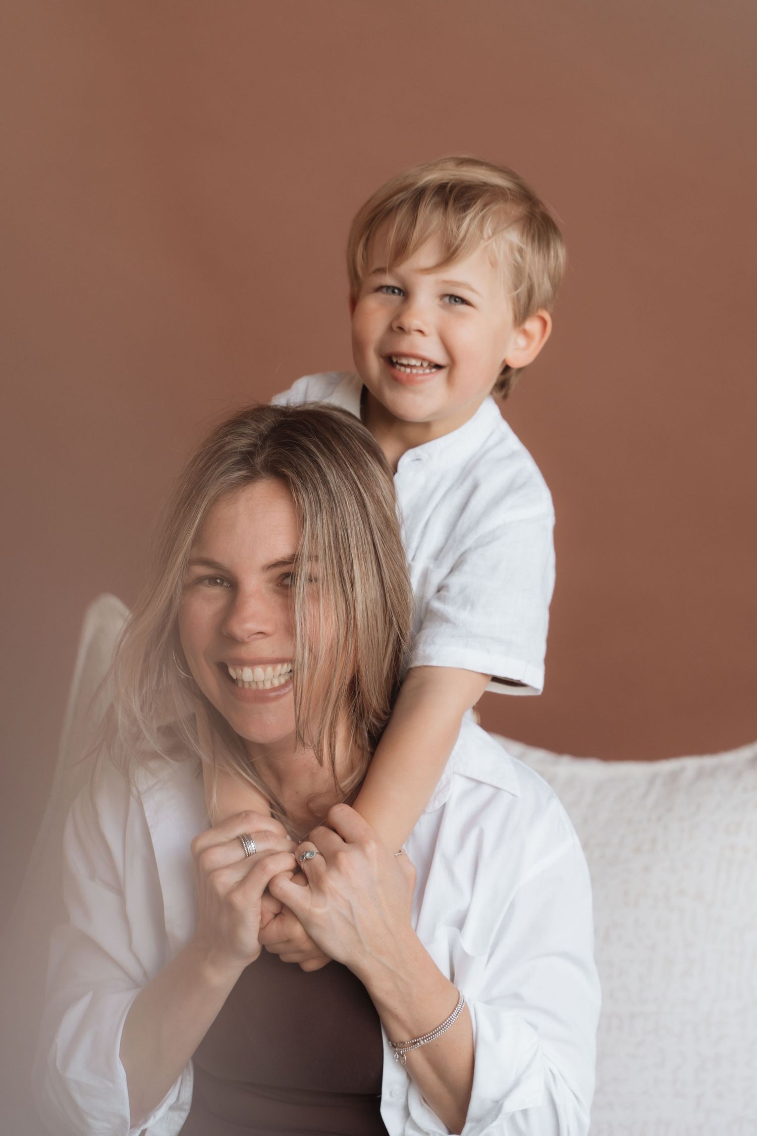 A smiling woman and young boy in white shirts share a joyful piggyback moment against a warm brown background.