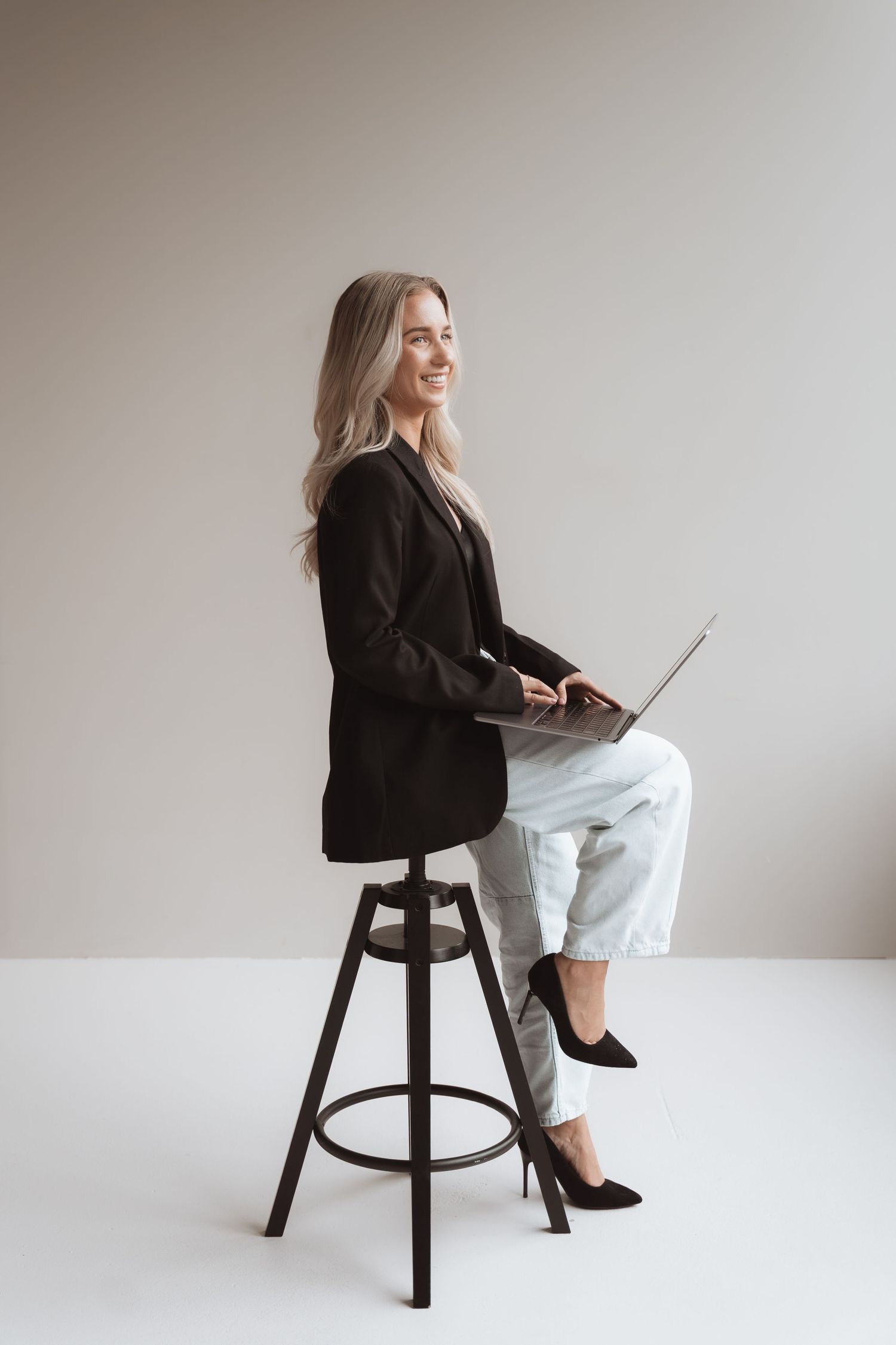 Smiling blonde businesswoman sitting on stool with laptop, wearing black blazer, jeans, and heels against gray background.