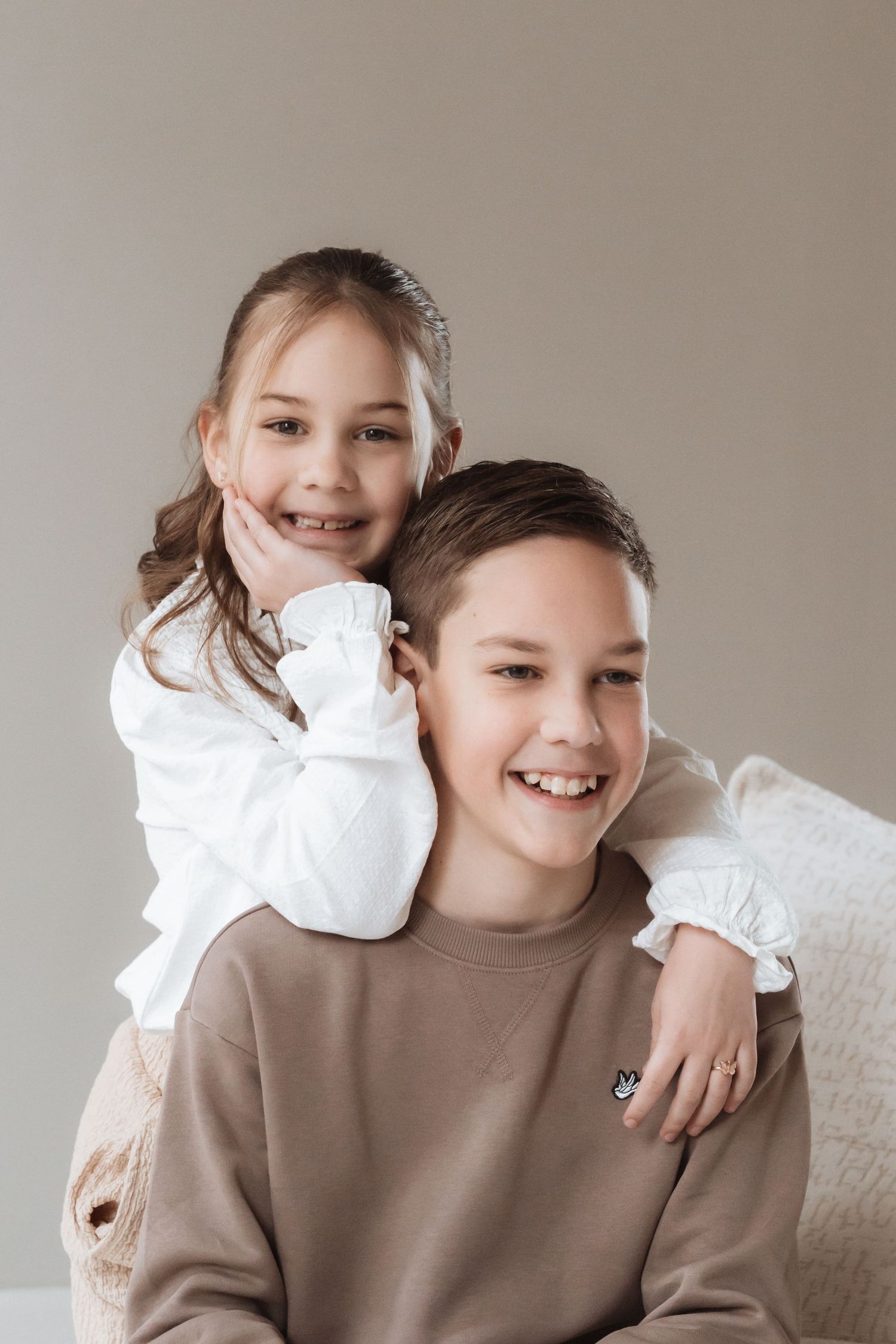 A smiling boy in a beige sweatshirt with a younger girl in a white blouse hugging him from behind against a gray background.