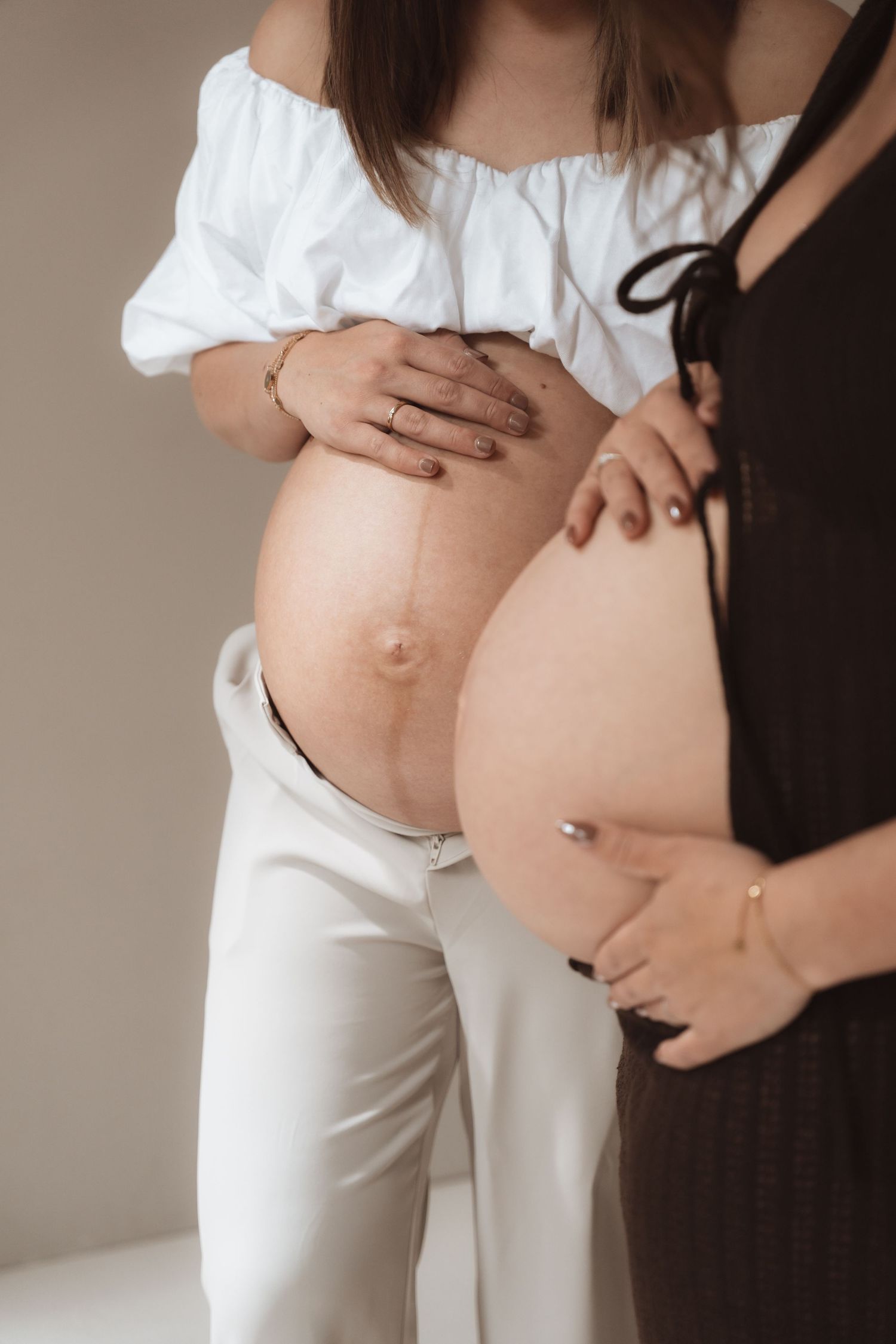 Two pregnant women standing side by side, showcasing their baby bumps, one in white outfit and one in black.
