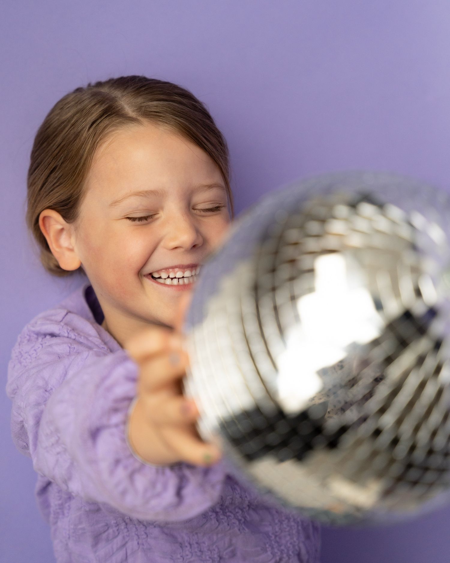 Child in purple sweater joyfully playing with a silver disco ball against purple background.