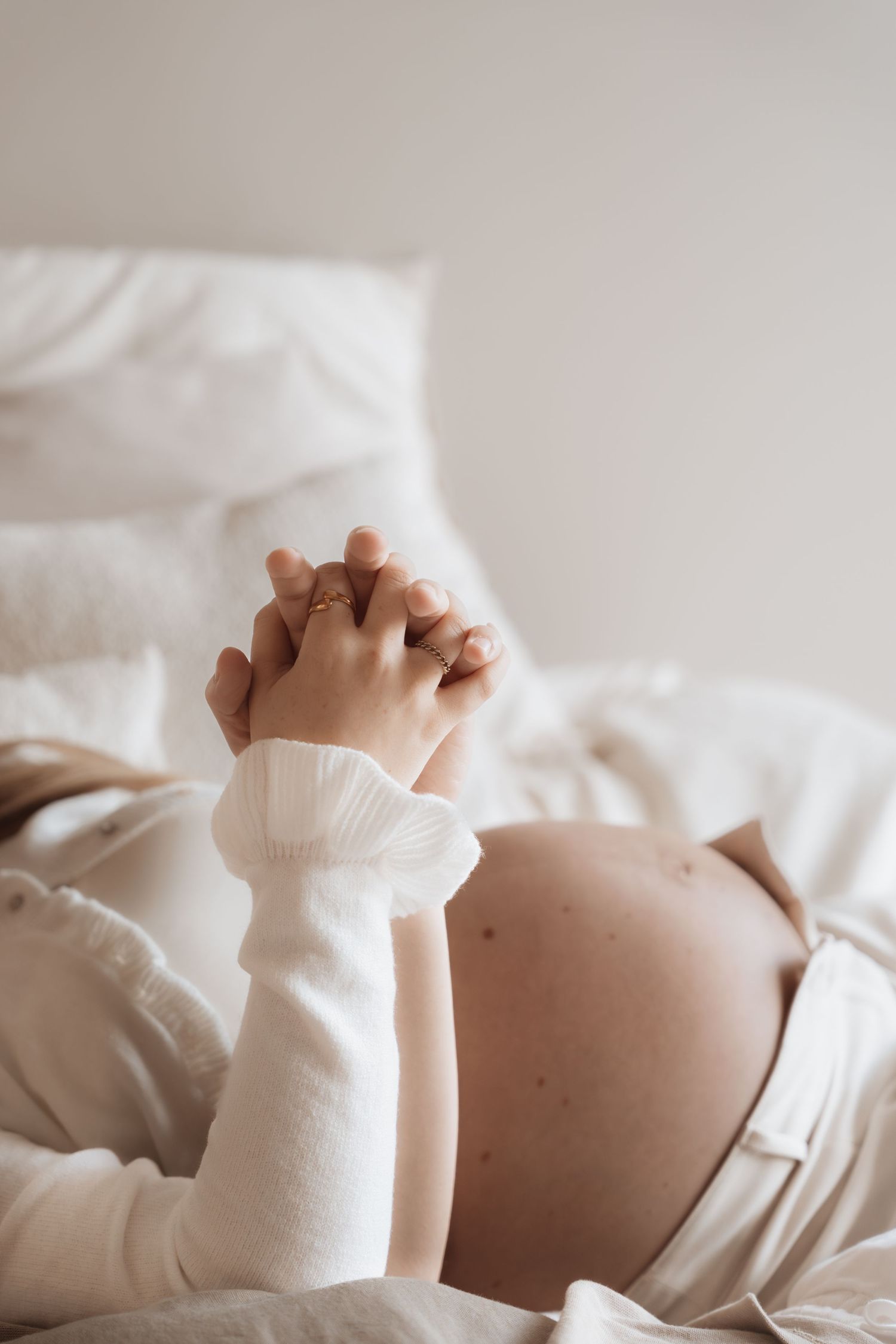 Pregnant woman lying on white bed with hands clasped over baby bump, wearing cozy white sweater and gold rings.