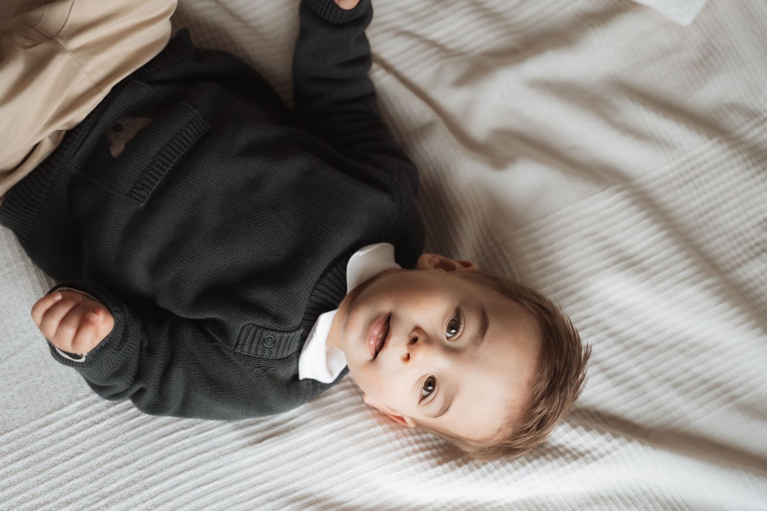 Small child wearing a dark hoodie lies on striped bedding while looking up at the camera.