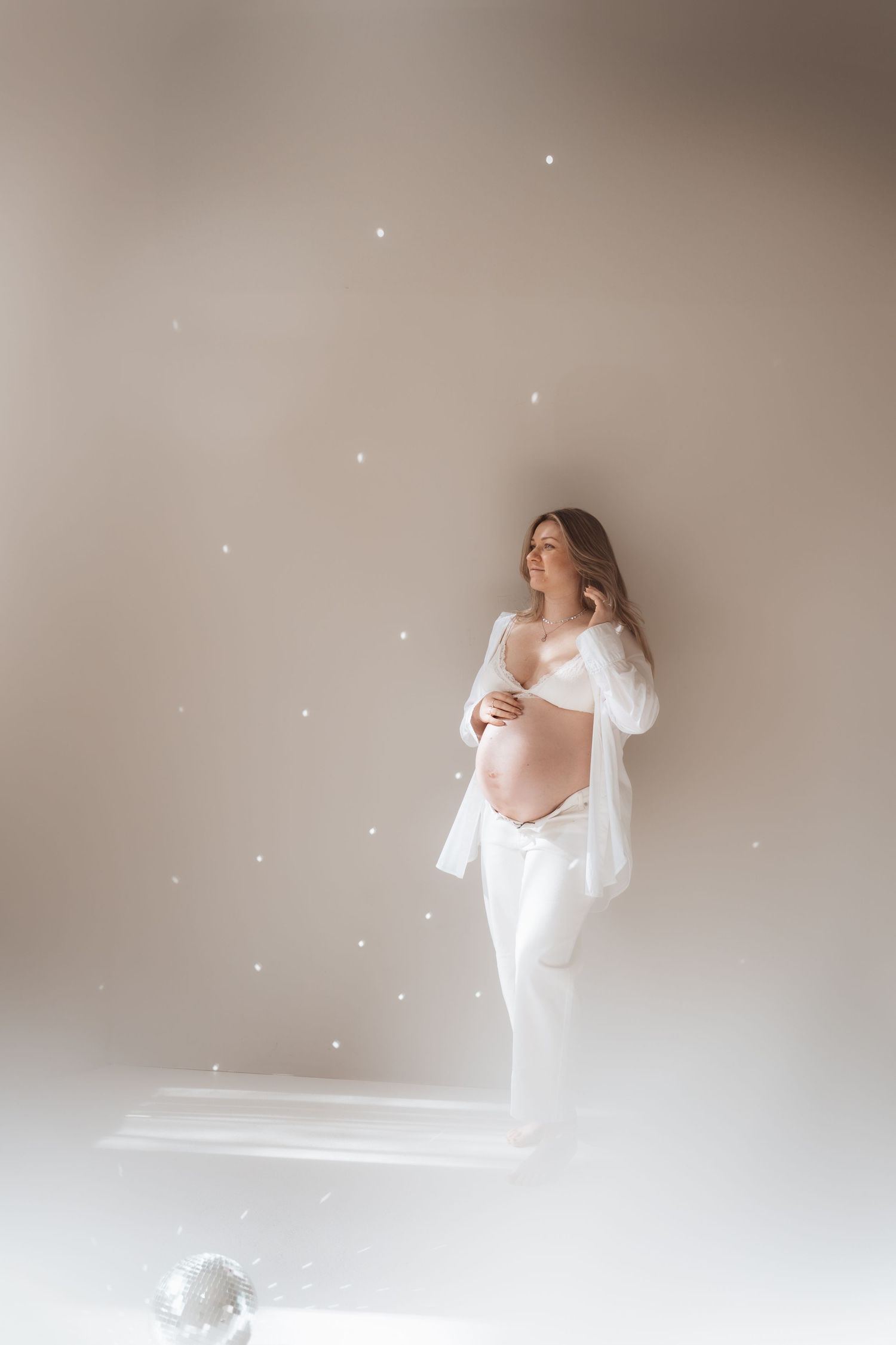 Pregnant woman in white outfit cradling baby bump, surrounded by sparkling lights in a dreamy studio setting.