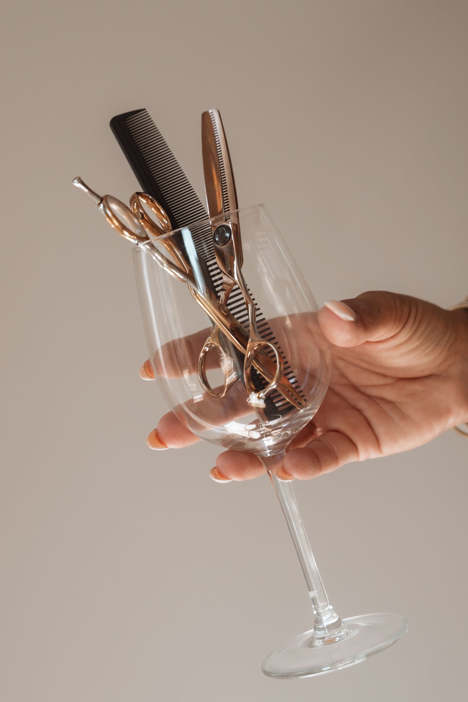 A hand holds a wine glass containing gold scissors and a black comb against a neutral background.