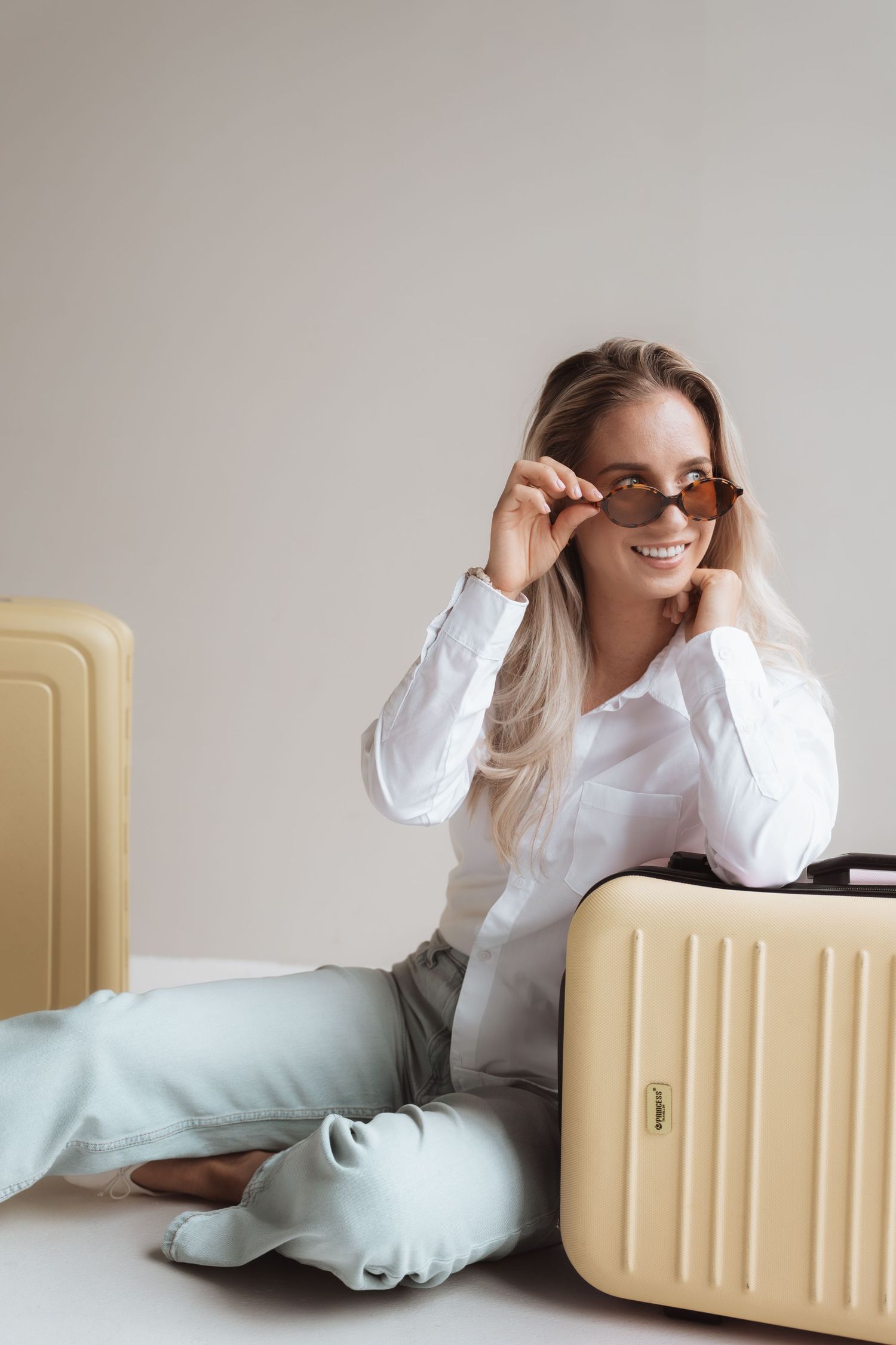 Smiling woman in white shirt adjusting sunglasses, sitting between yellow suitcases on white background.