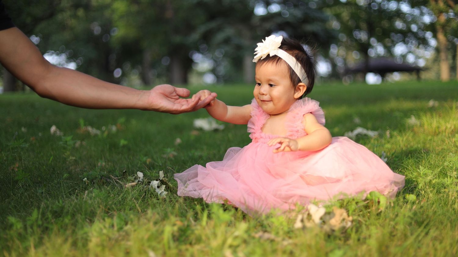 Toddler girl in pink dress reaching toward parent’s hand during outdoor portrait session