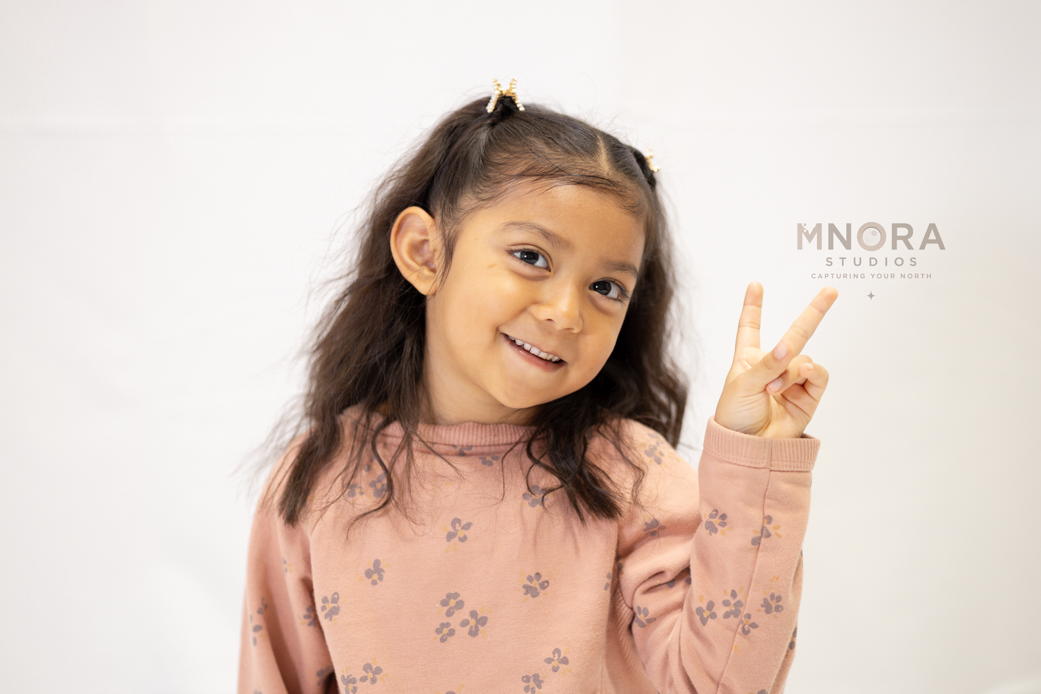 Girl in pink sweater posing with peace sign in front of MNORA Studios backdrop