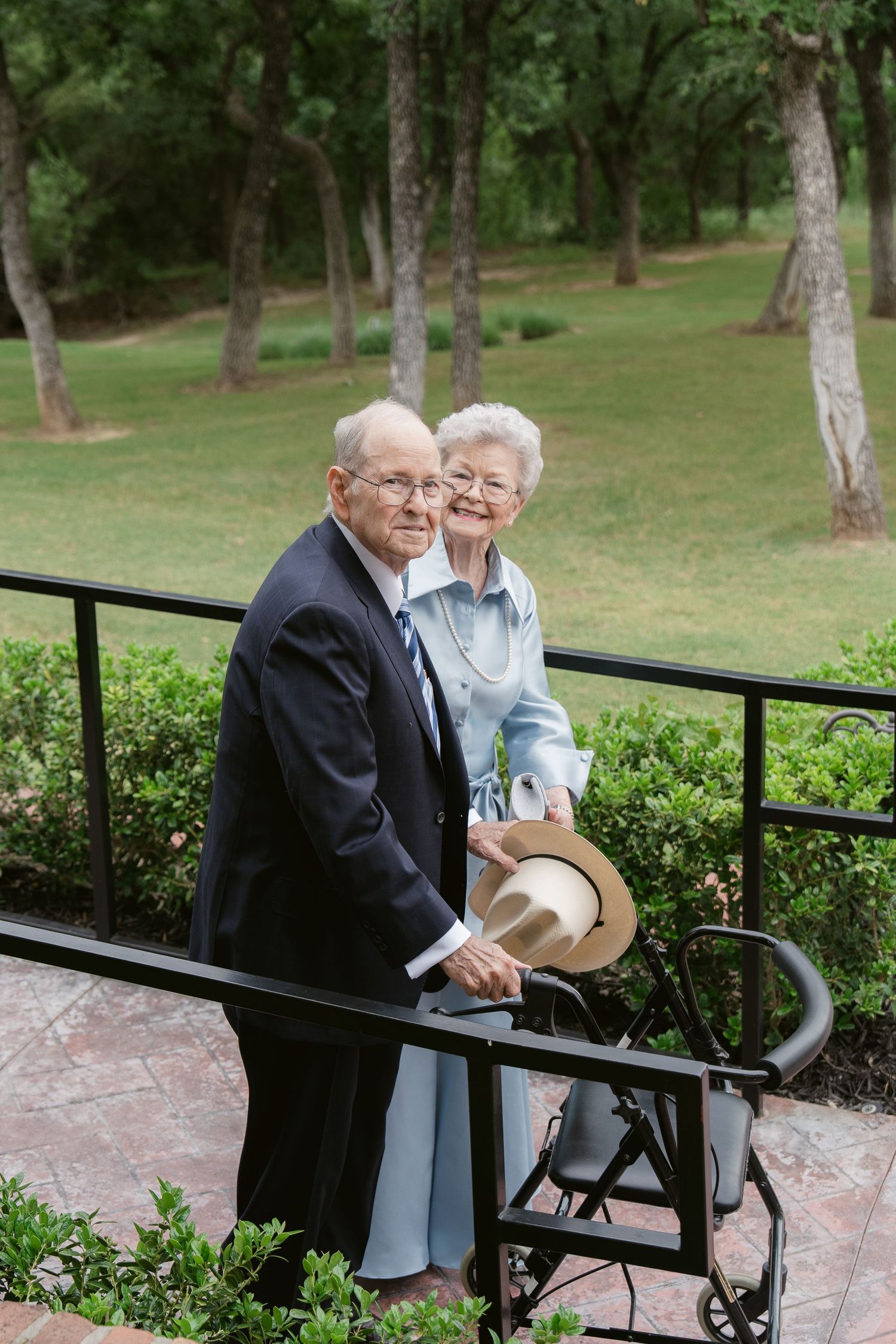 Elderly couple posing outdoors on a patio, man in dark suit holding hat, woman in light blue dress, lush green trees behind.