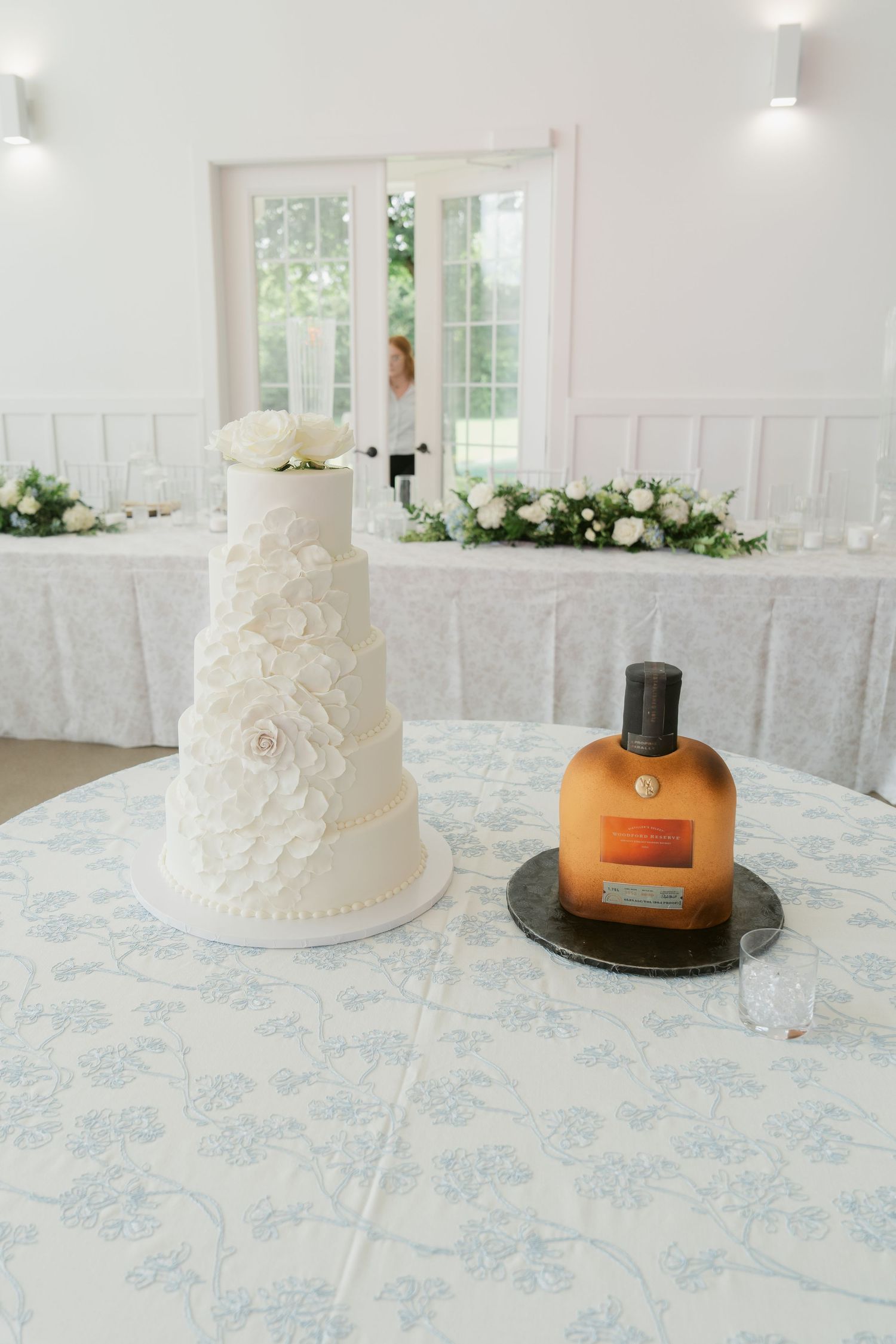 Elegant four-tier white wedding cake with floral details beside a perfume bottle cake on a floral tablecloth.