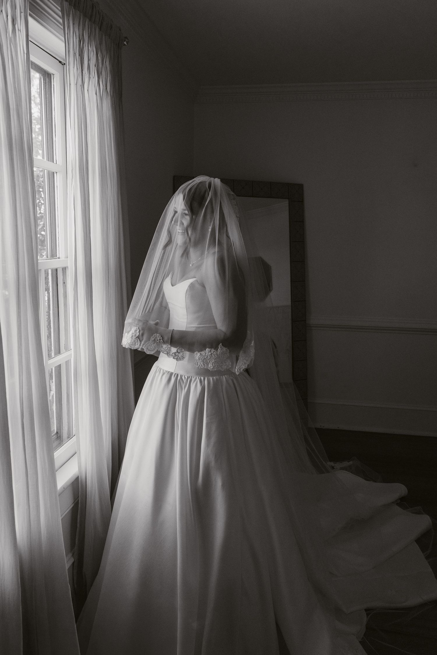 Black and white photo of a bride in a white gown and veil standing by a window in a softly lit room.