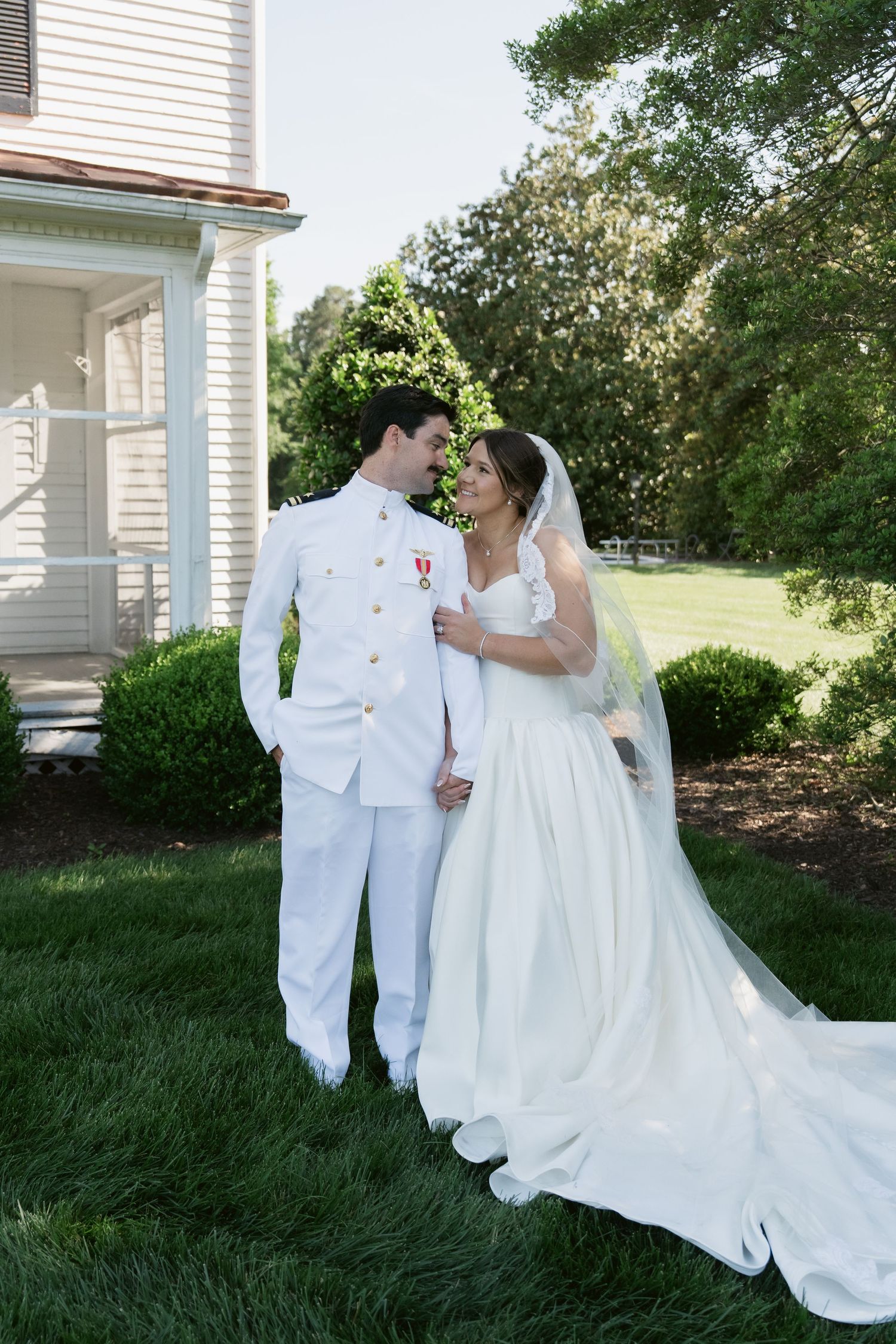 Bride and groom in white wedding attire stand together on green lawn outside white house, sharing a romantic moment.