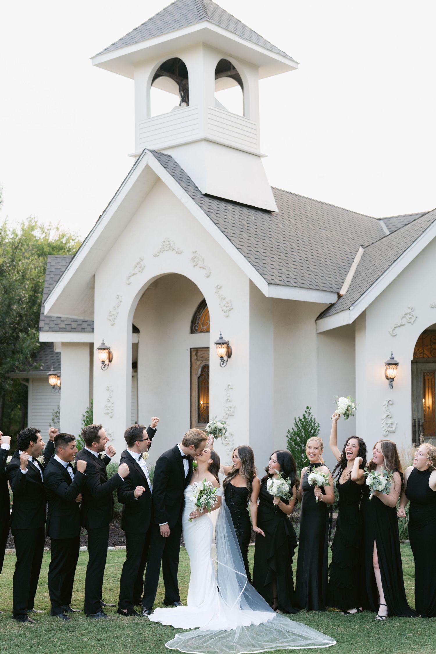 Wedding party in black attire poses outside white chapel as bride and groom share a kiss.